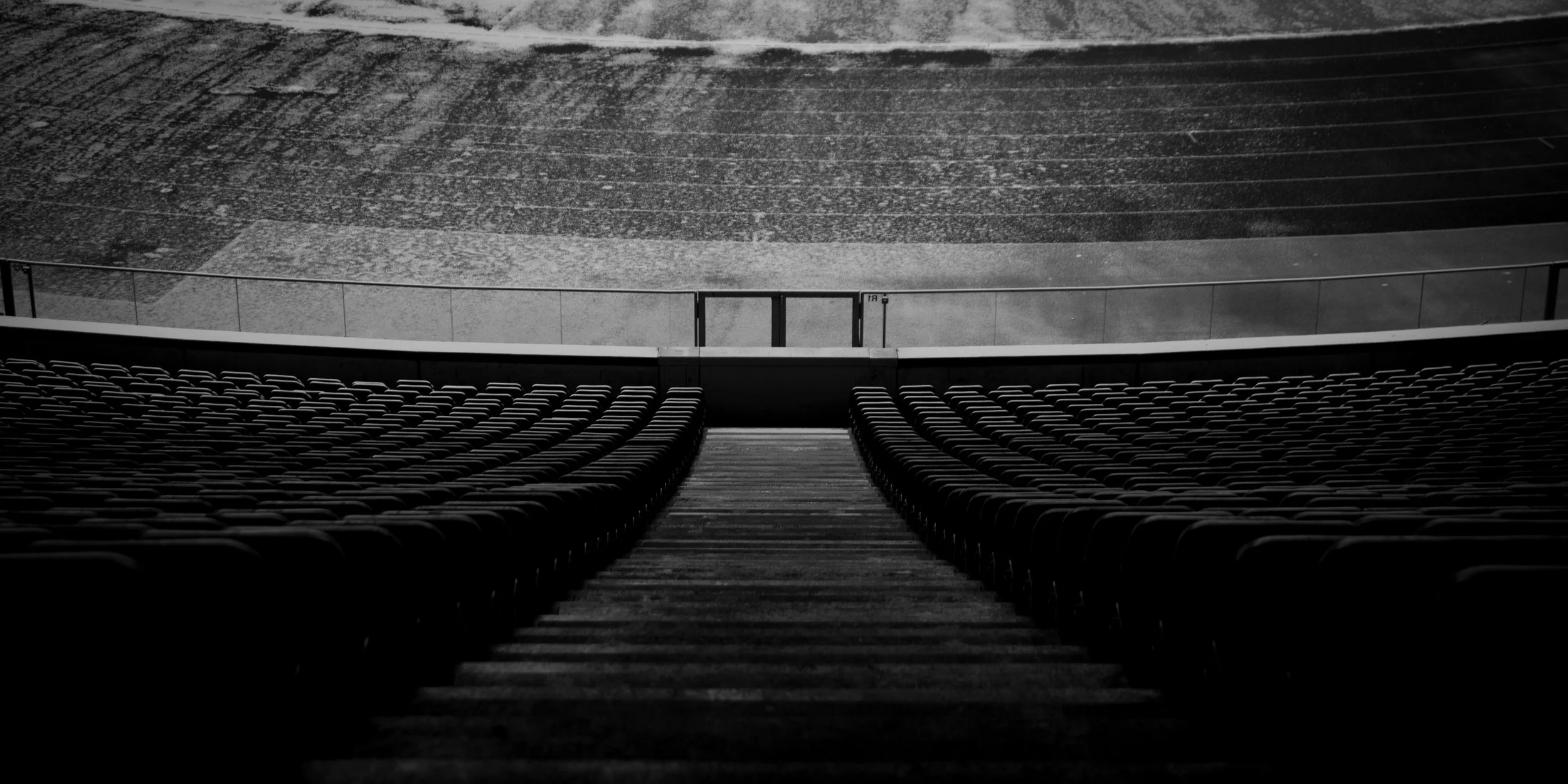 Empty stadium with rows of seats, stairs leading to a railing, and a playing field visible in the background.