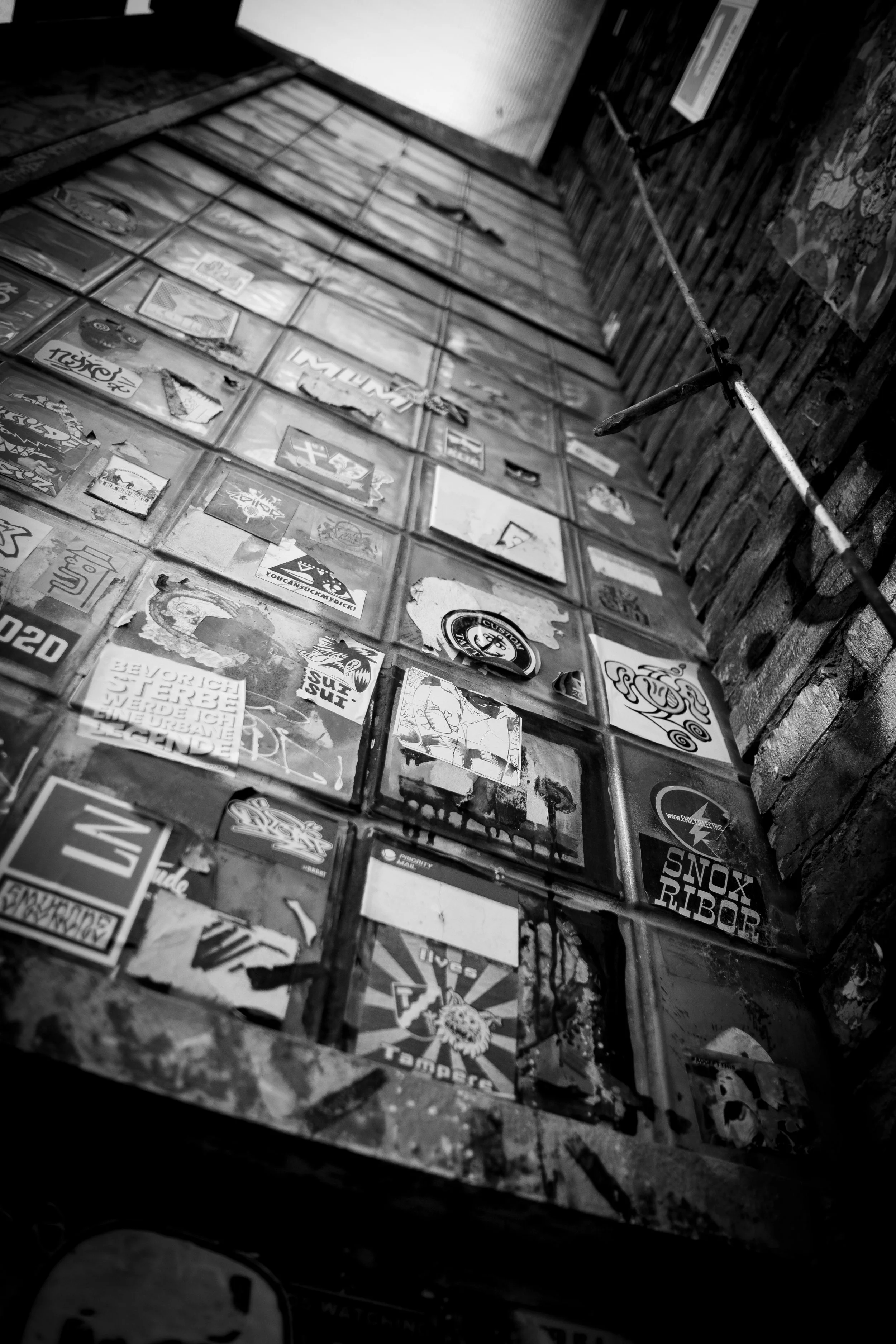 A black and white photo of a tall wall covered with numerous stickers, with a brick wall on the right side and a pipe running diagonally across the image.