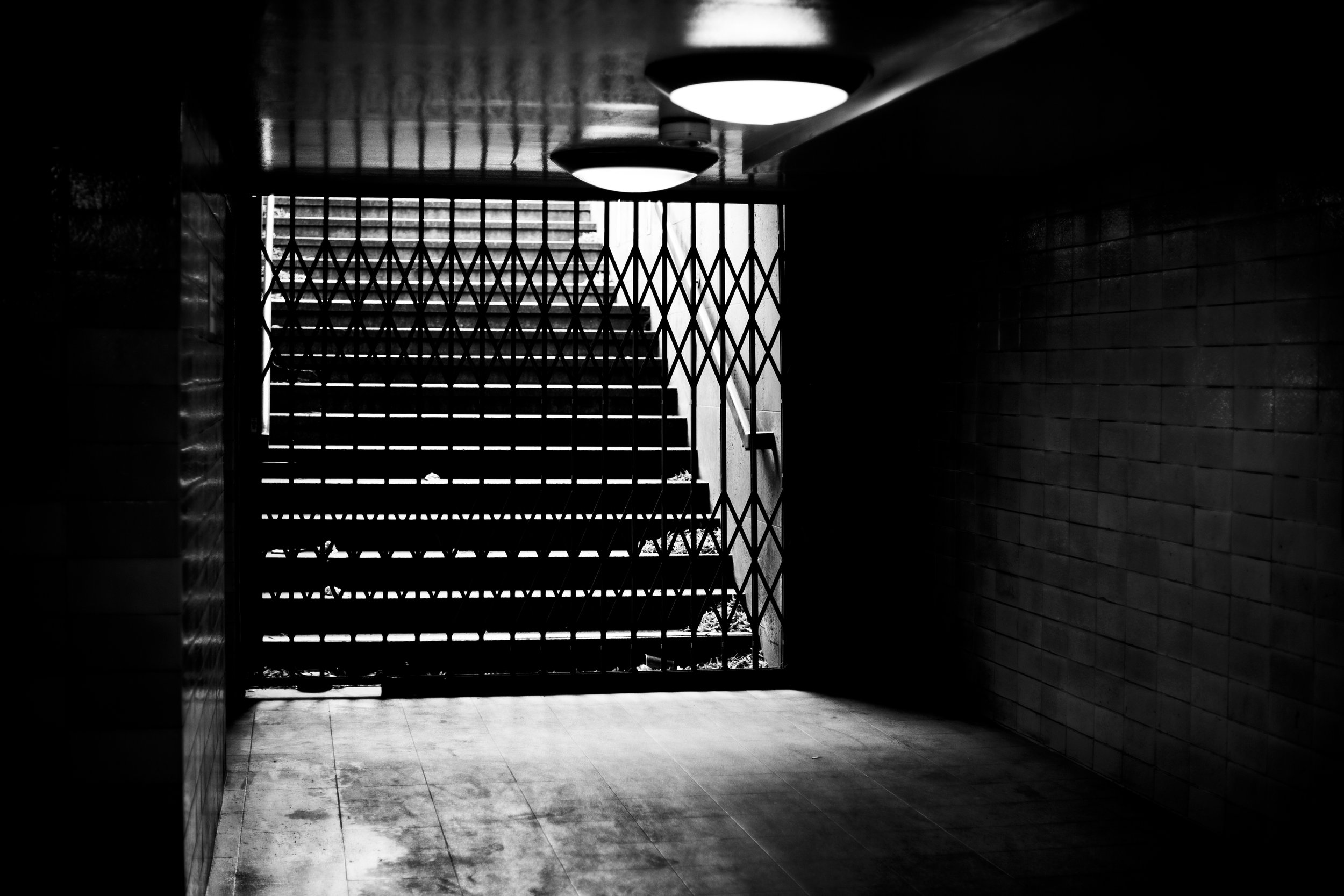 Black and white photo of a staircase behind a metal gate, with the light from outside illuminating part of the scene. Ceiling lights are visible on the top, and the floor appears worn.