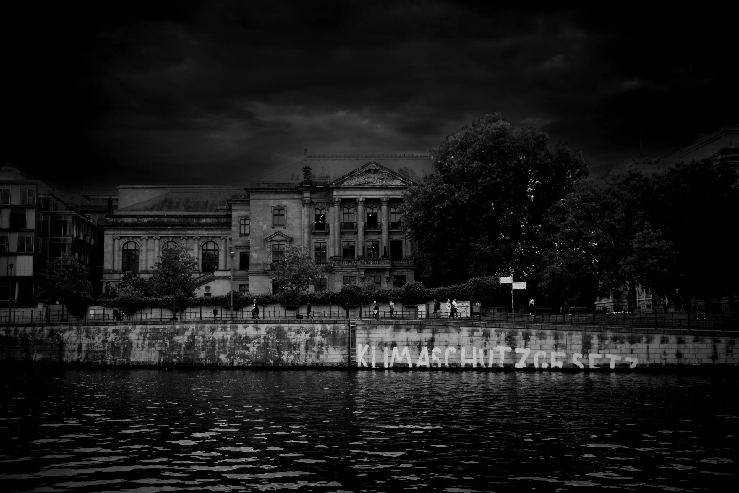 A black and white photograph of a historic building along a river with graffiti on the wall that reads 'Klimaschutzgesetz'. There are trees and pedestrians in front of the building, and dark clouds overhead.
