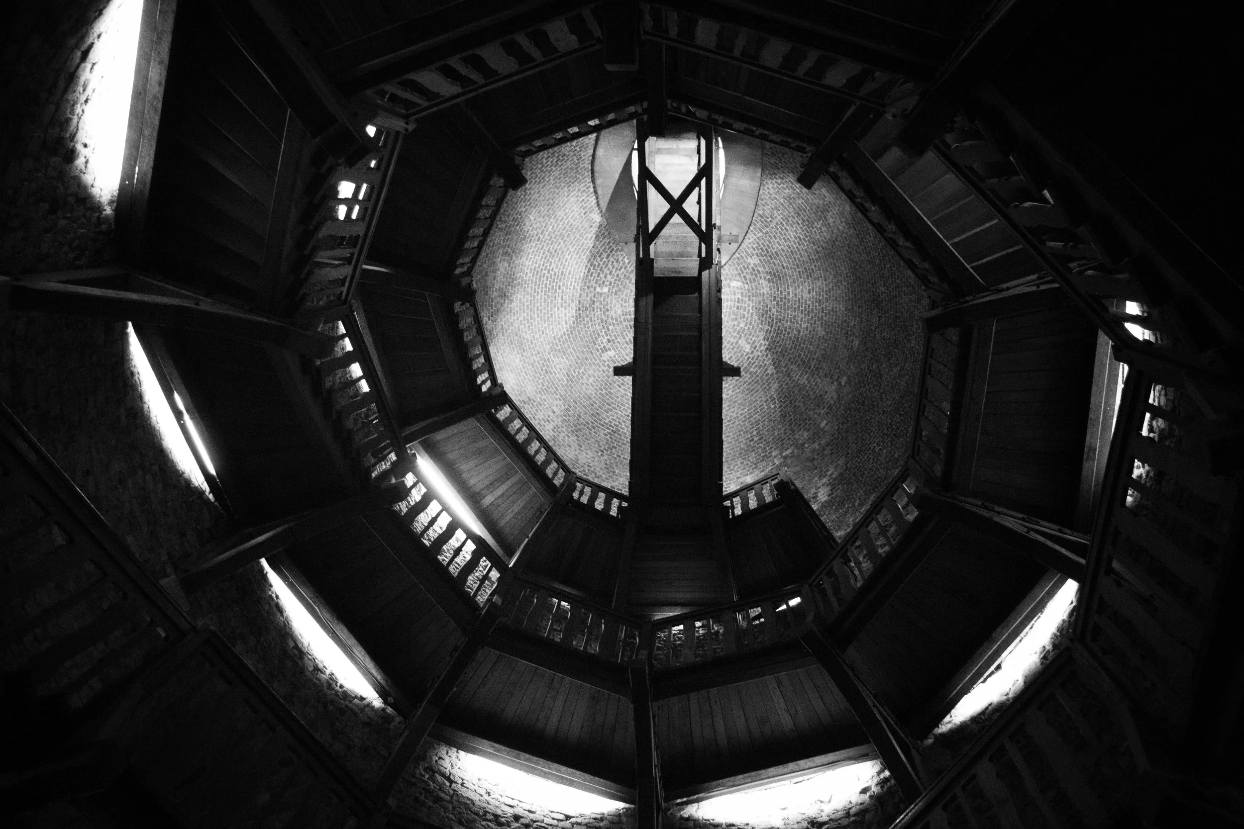 View looking up at an octagonal wooden staircase with railings, leading to a circular brick courtyard below, in black and white.