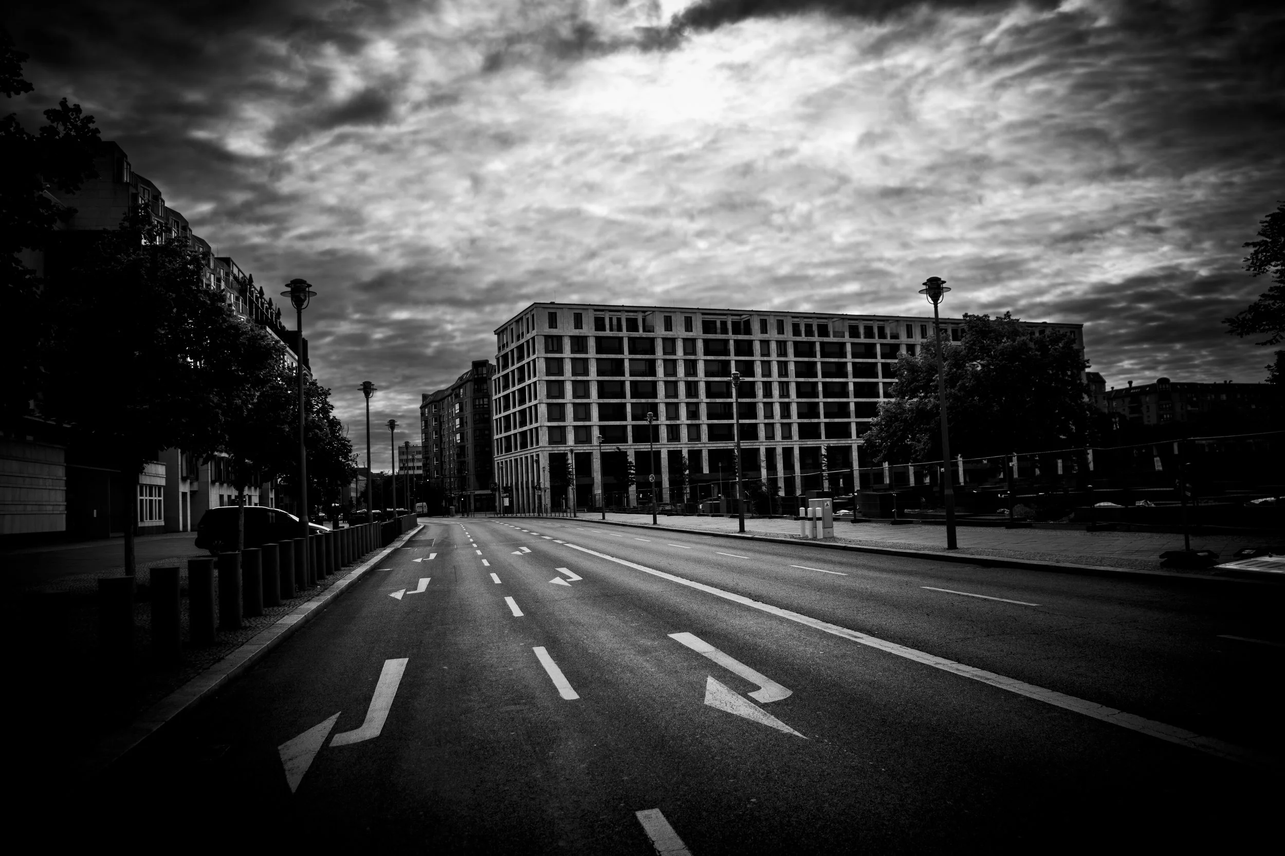 Empty city street in black and white with buildings, streetlights, and cloudy sky.
