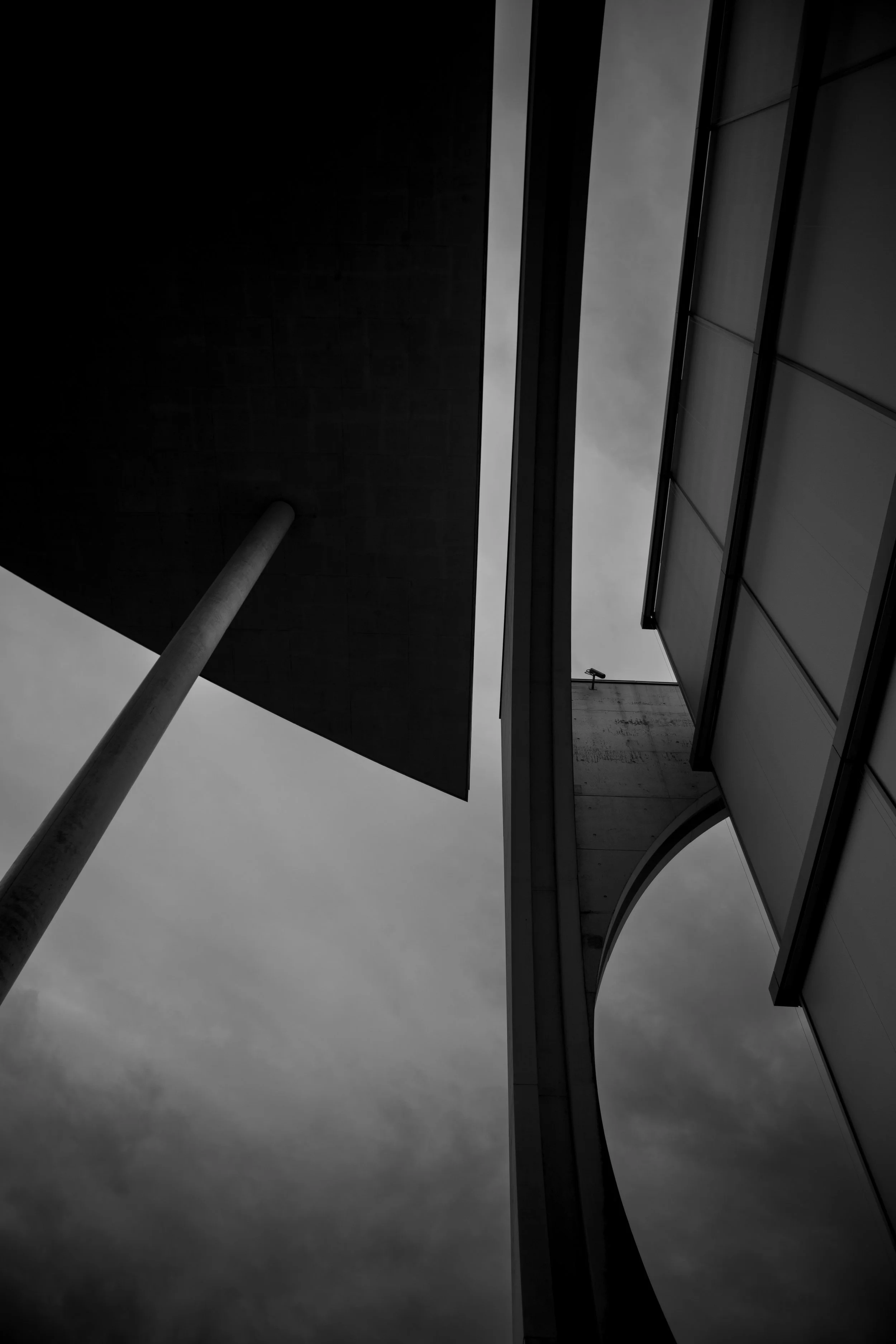 Low-angle black-and-white photograph of modern architectural buildings against cloudy sky, featuring a large supporting pole and geometric shapes.