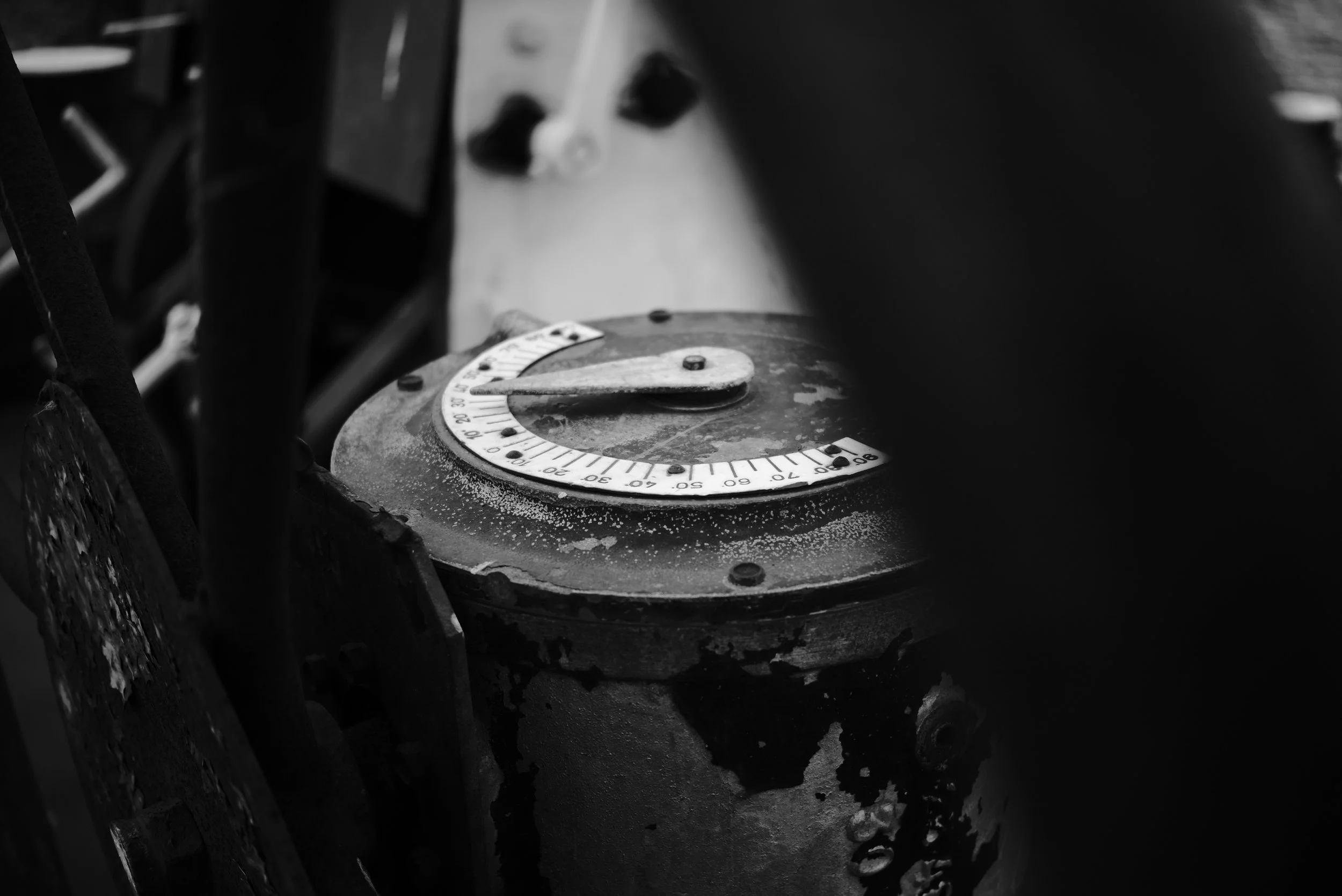 A close-up of an industrial or mechanical control dial with a pointer, mounted on a worn, weathered surface, with other machinery parts in the background.