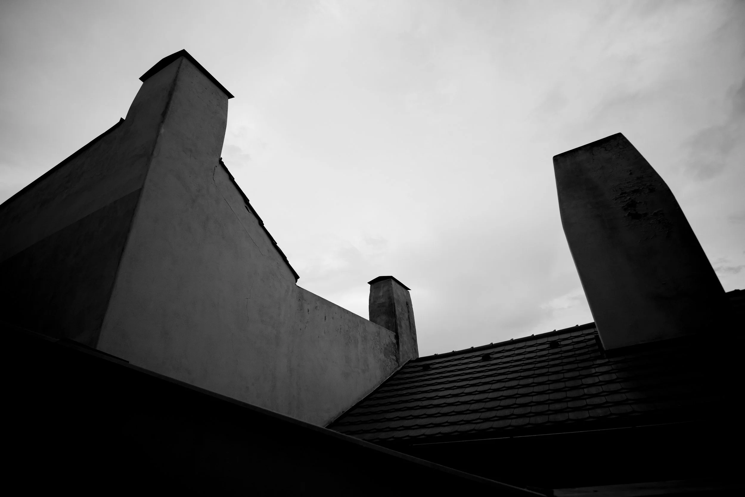 Black and white photo of rooftops with three chimney stacks against a cloudy sky.