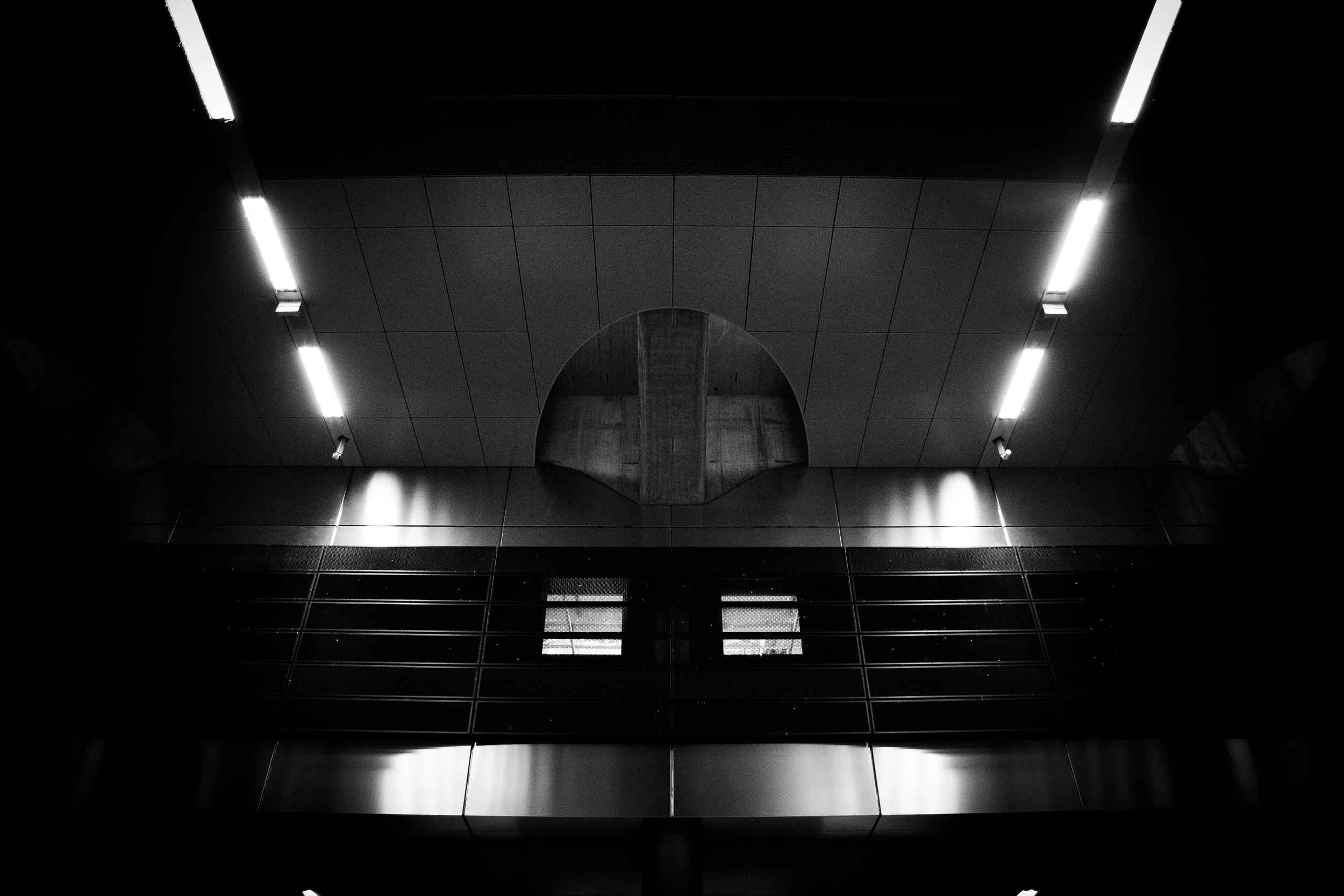 Black and white photo of modern building interior, displaying ceiling with bright light fixtures, a circular opening, and a reflective glass surface.