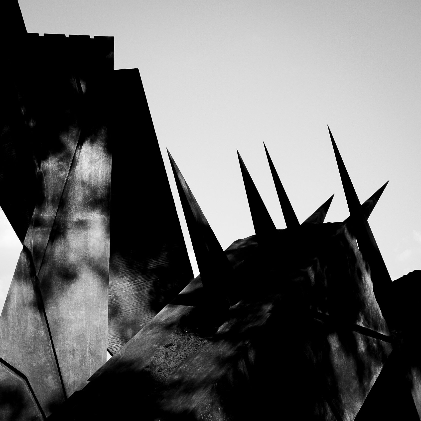 Black and white photograph of a modern sculpture with sharp, spiked elements and metallic surfaces, set against a sky with a few clouds.