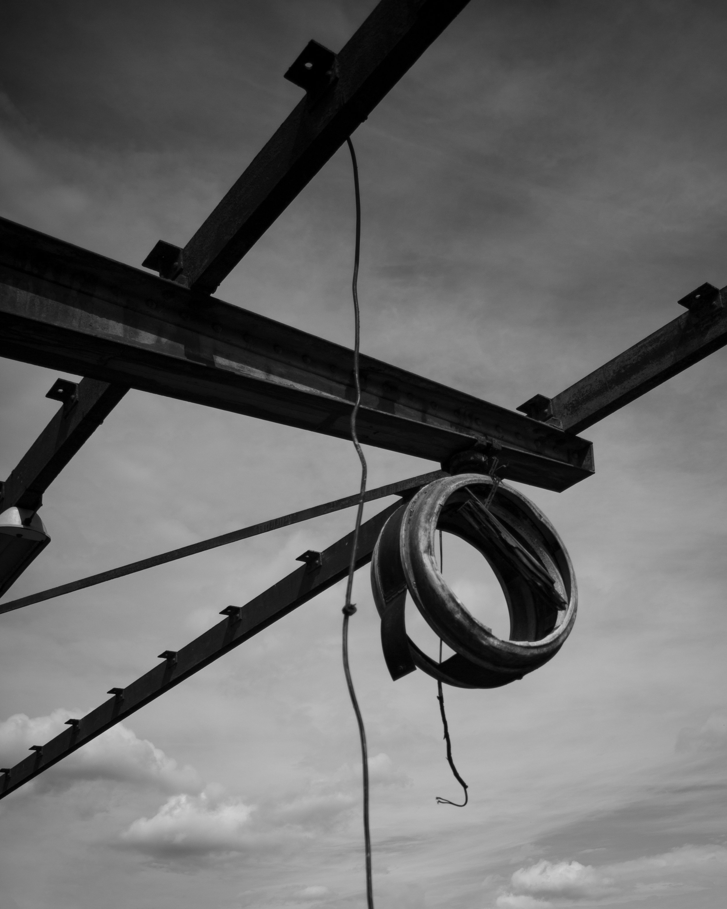 Black and white photo of a construction site with metal beams and a large metal ring hanging from a wire against a cloudy sky.