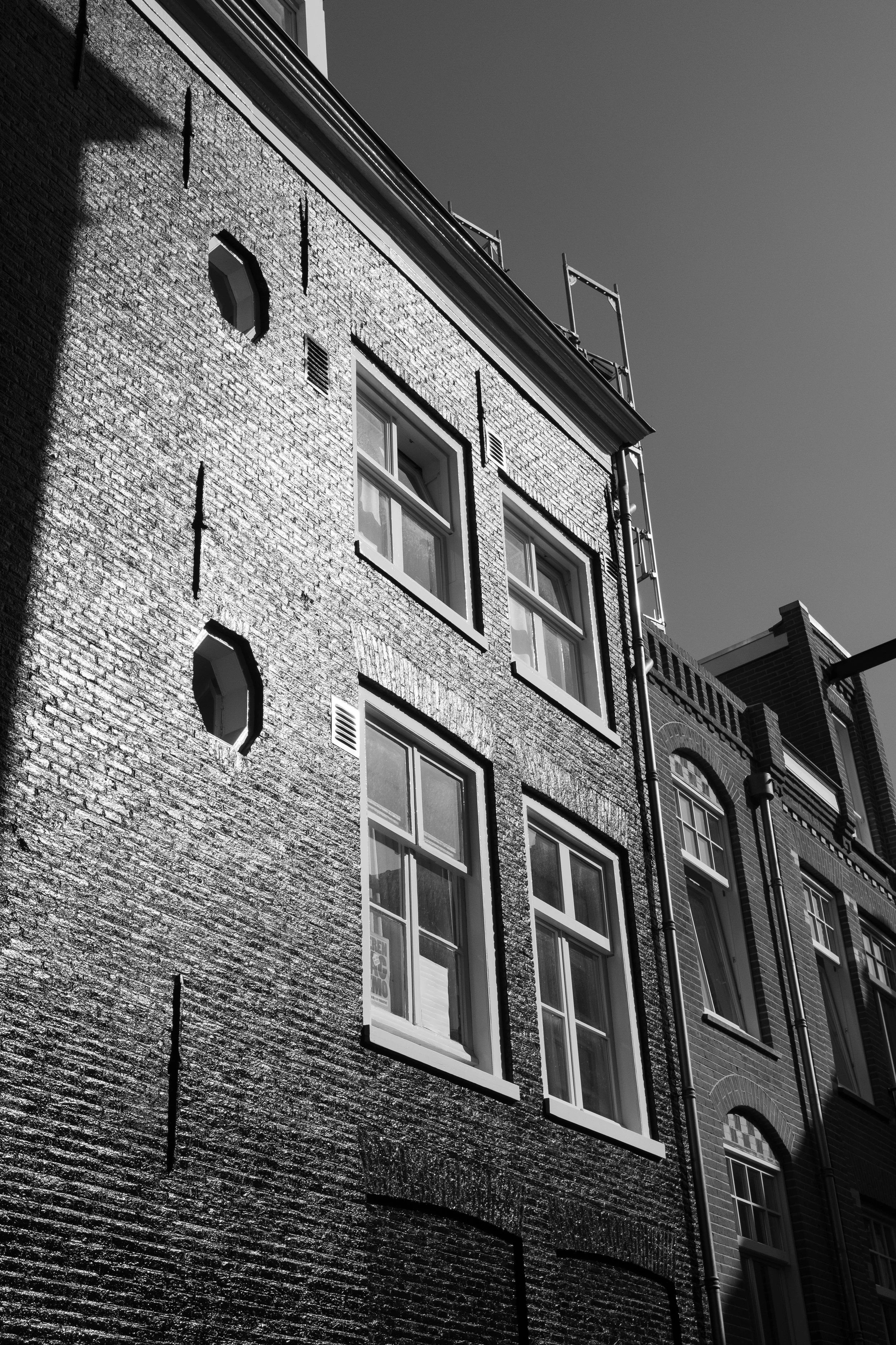 Black and white photograph of a brick building with three windows, shadows cast on the exterior wall, and scaffolding on the side against a clear sky.