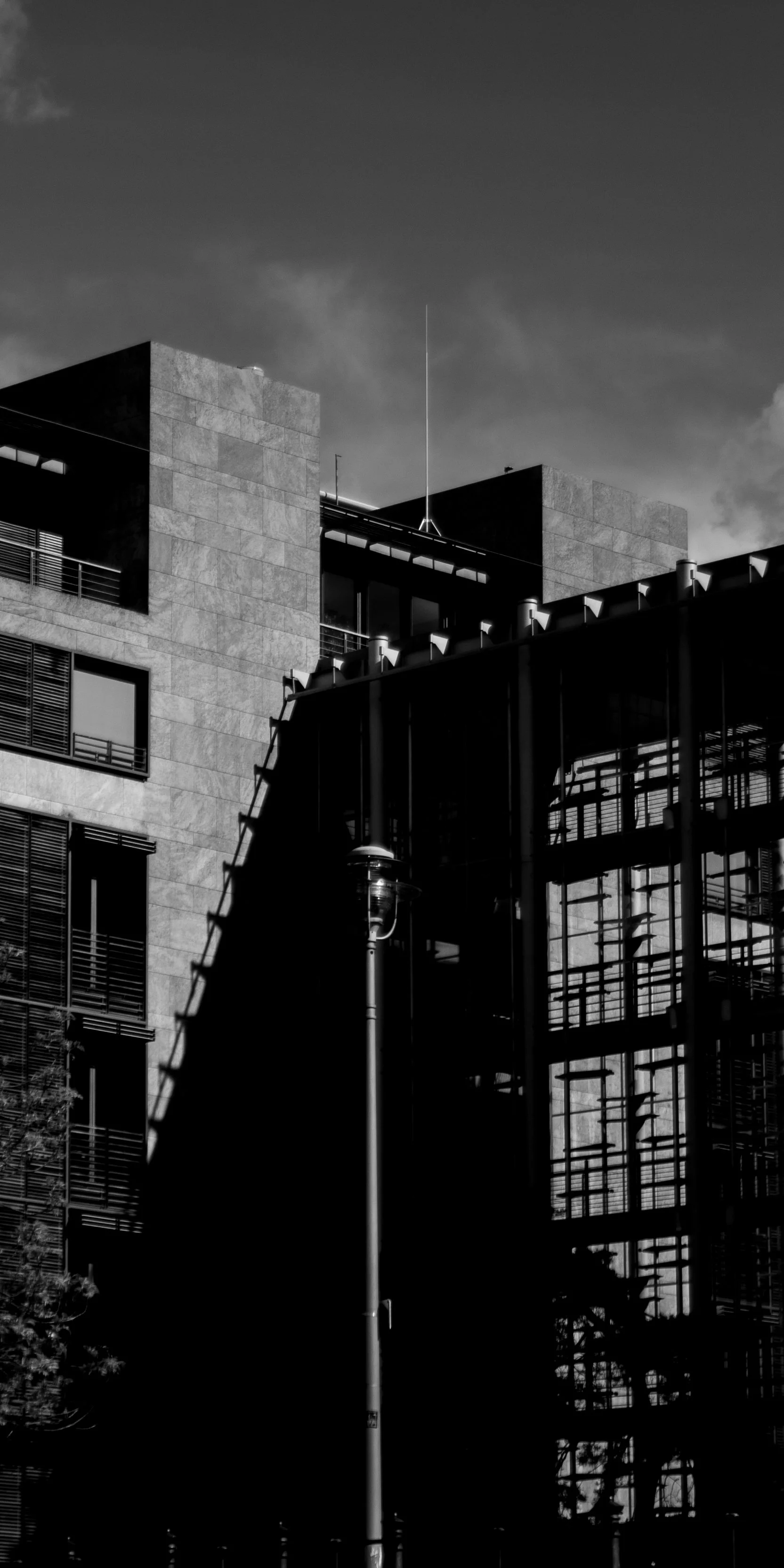 Black and white photo of modern buildings with a lamp post in the foreground and a cloudy sky above.