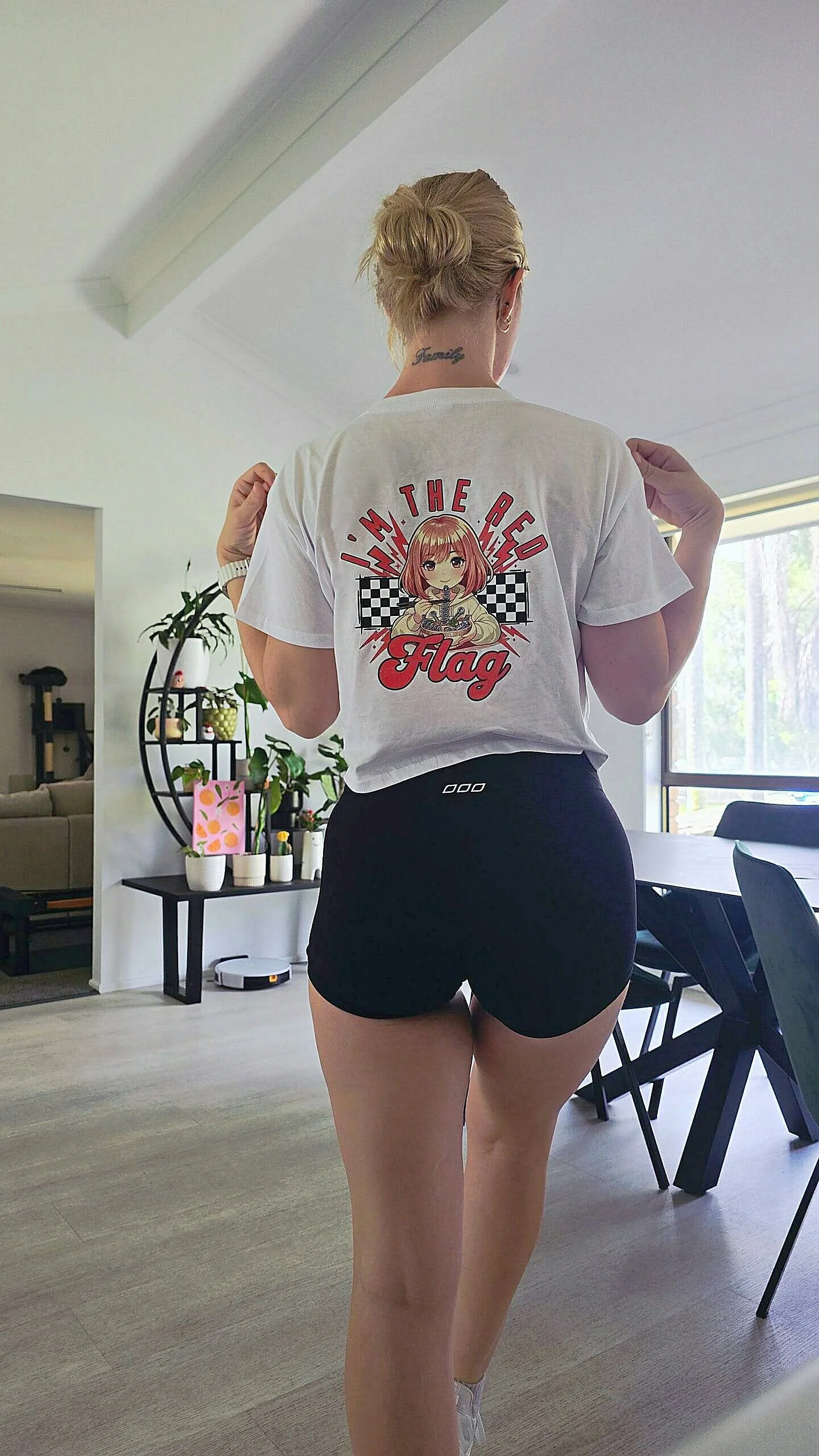 A woman standing in a living room with her back facing the camera, wearing a white cropped T-shirt with the graphic of a cartoon anime girl and the words 'I'm the red flag,' and black shorts.