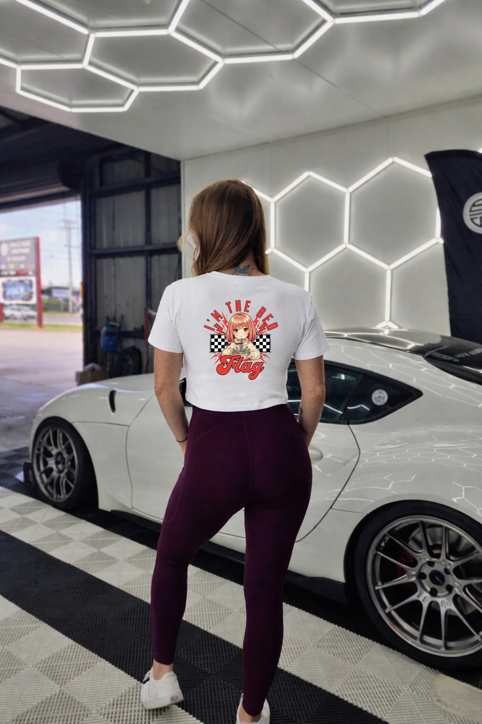 Back of a person with shoulder-length blonde hair wearing a white crop T-shirt with a graphic on the back, featuring a girl with anime graphic of a young girl, with checkered background, and text 'I'm the Red Flag' under a hexagon light garage standi