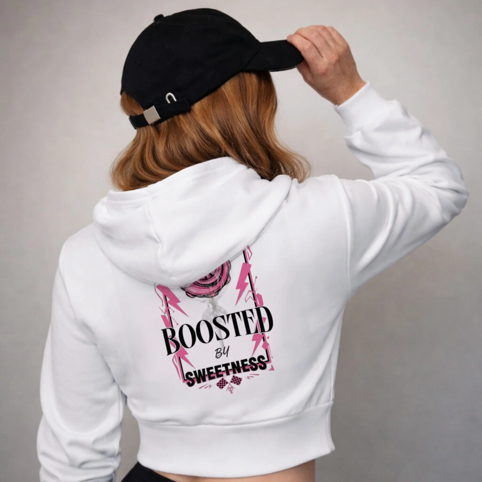 Back of person wearing a white cropped hoodie with pink and black text that reads "Boosted by Sweetness" and a black cap, with long light brown hair.