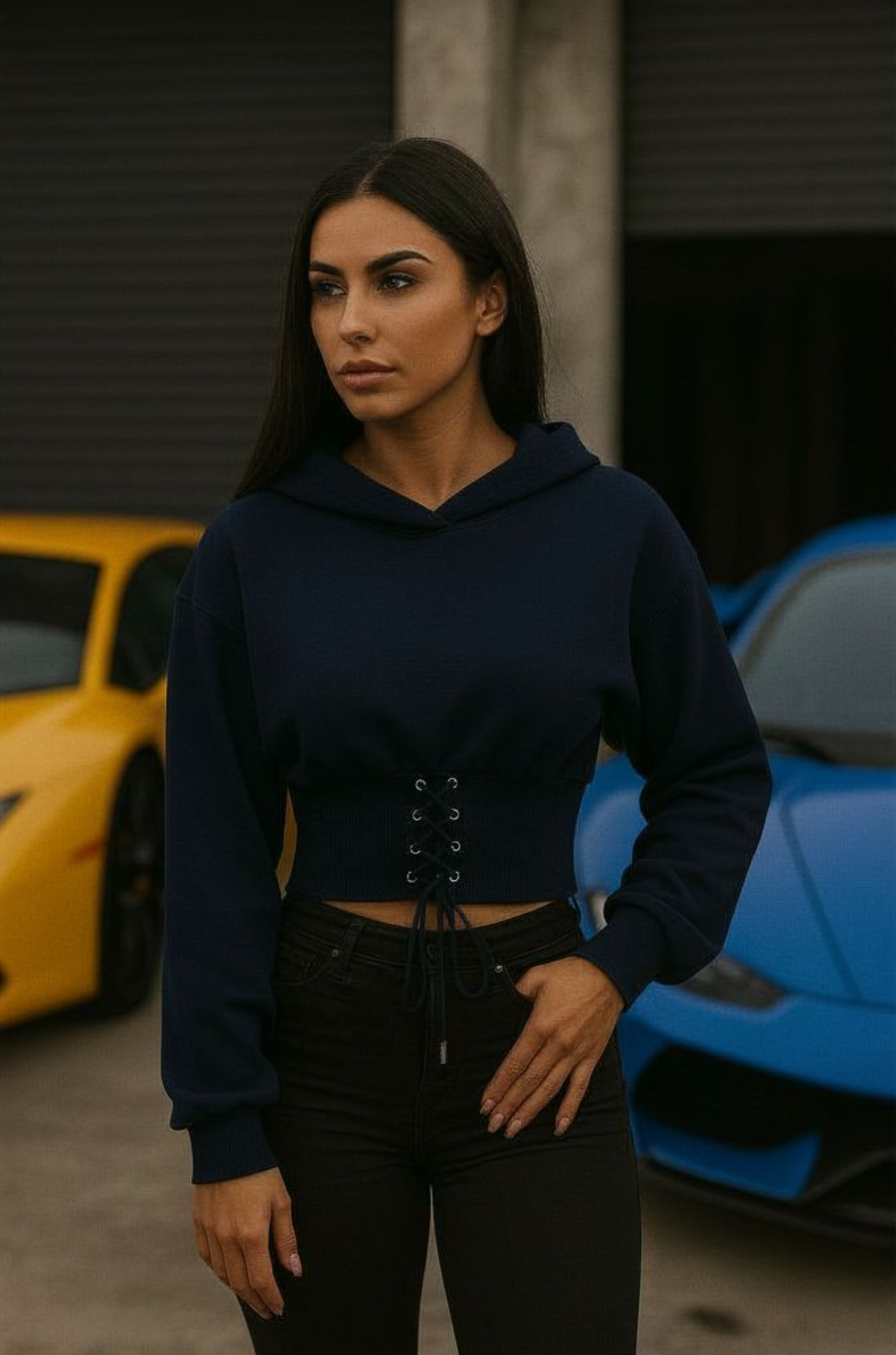 A woman standing in front of two sports cars, one yellow and one blue, wearing a navy crop top hoodie with lace-up details at the waist and black pants.