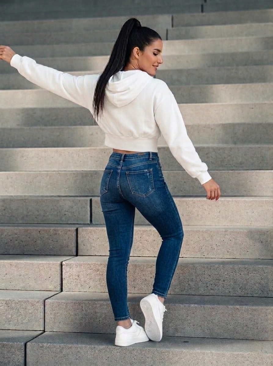 A woman with long dark hair up in a ponytail, wearing a white cropped hoodie with a front lace-up detail with layered waistband, with blue denim skinny jeans, standing on a set of stairs