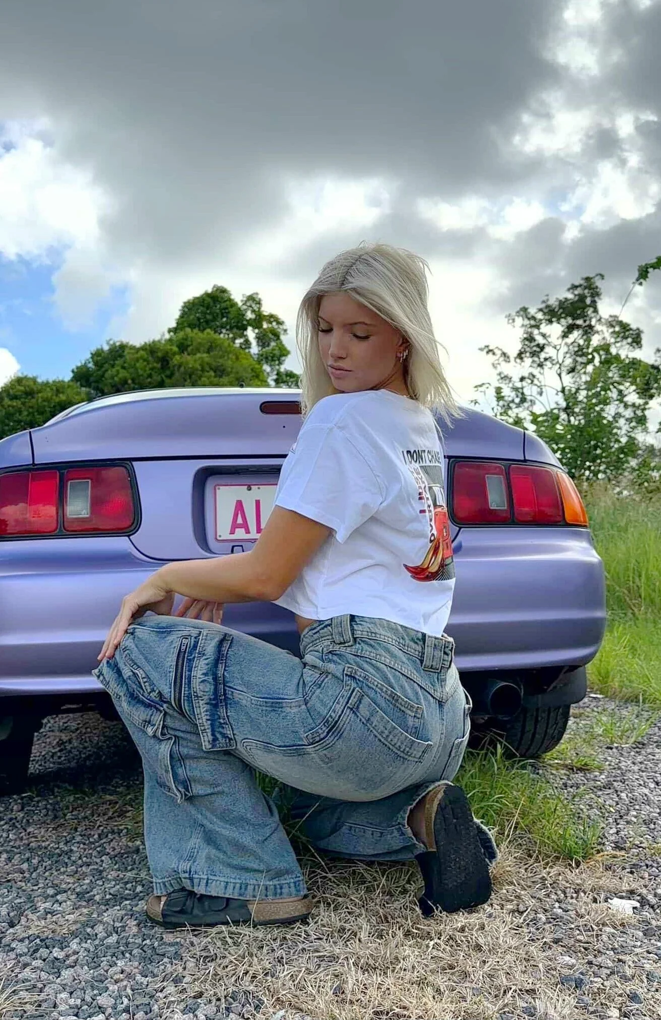 Young woman with blonde hair, wearing a white crop t-shirt with a red sports car graphic and the text 'I DON'T CHASE...I JUST OVERTAKE' on her back, crouching outdoors in front of her carl, with a baggy denim jeans.