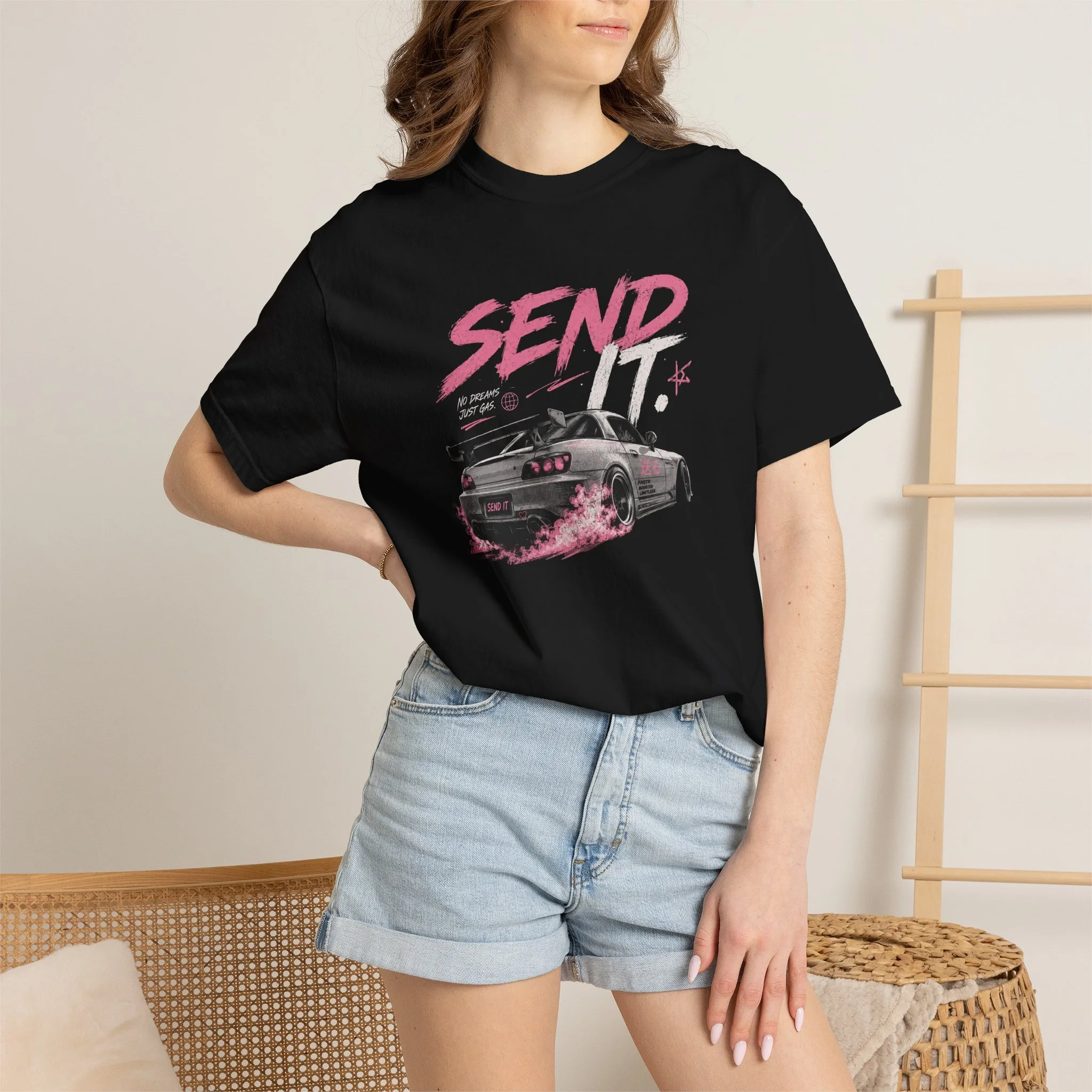 A woman wearing a black DRS oversized T-shirt with pink and white text that say "Send It', paired with denim shorts. The background features a woven chair and a wooden ladder-like decor piece on the wall.