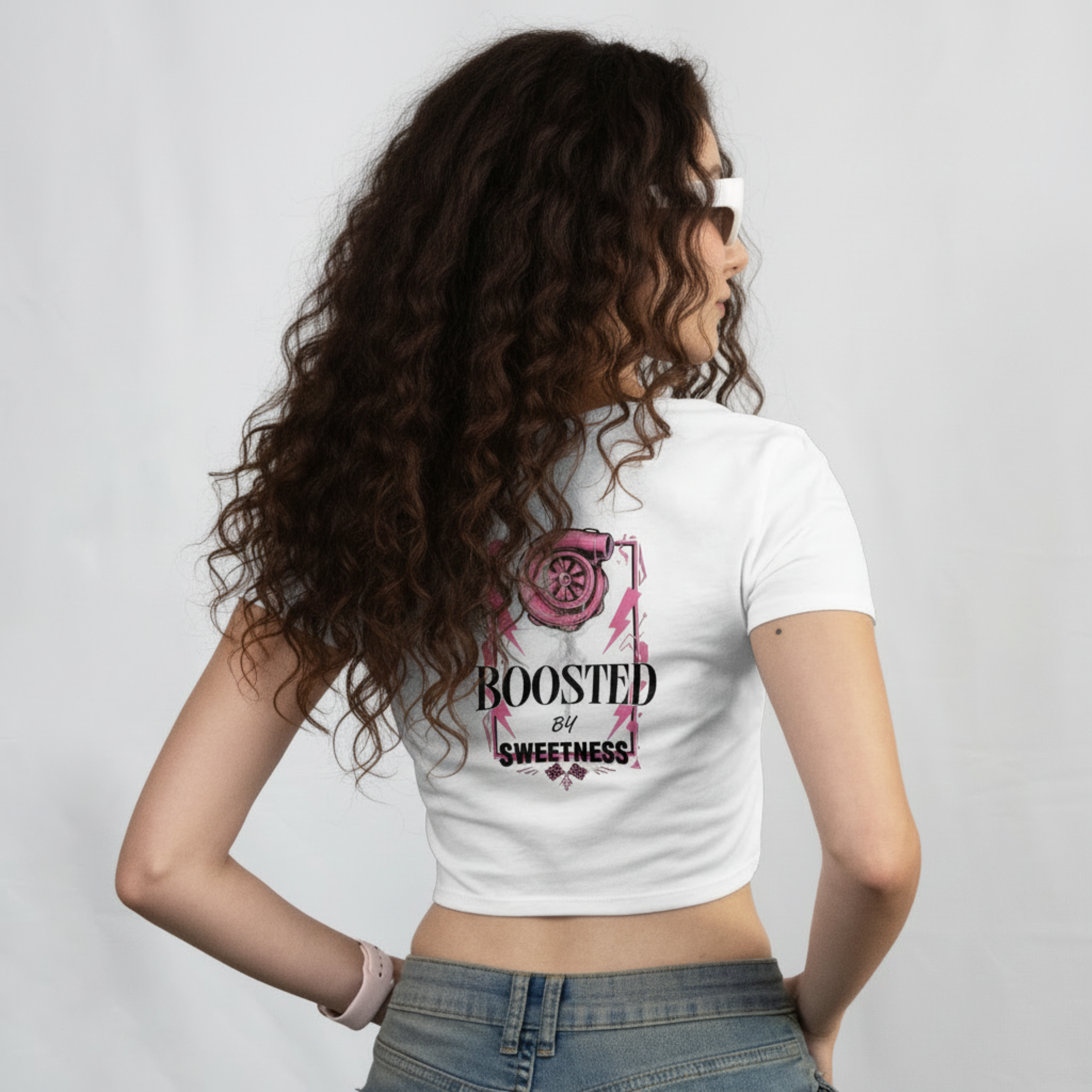 Woman with long curly hair wearing sunglasses and a white crop top that has ' Boosted by Sweetness' printed on the back with pink graphic design of a combination of which a turbine is the lollipop, standing against a plain background.