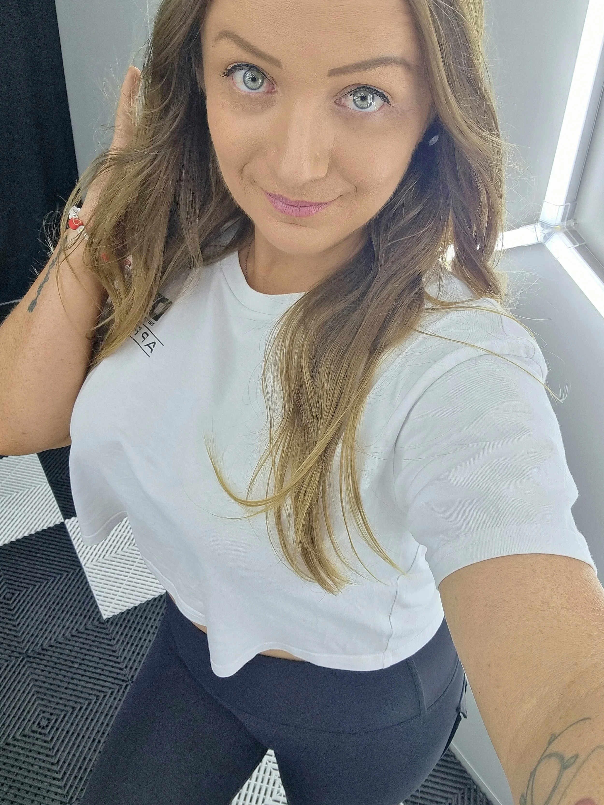A woman with long light brown hair and blue eyes taking a selfie indoors, wearing a DRS Apparel white t-shirt and black pants, near a window.
