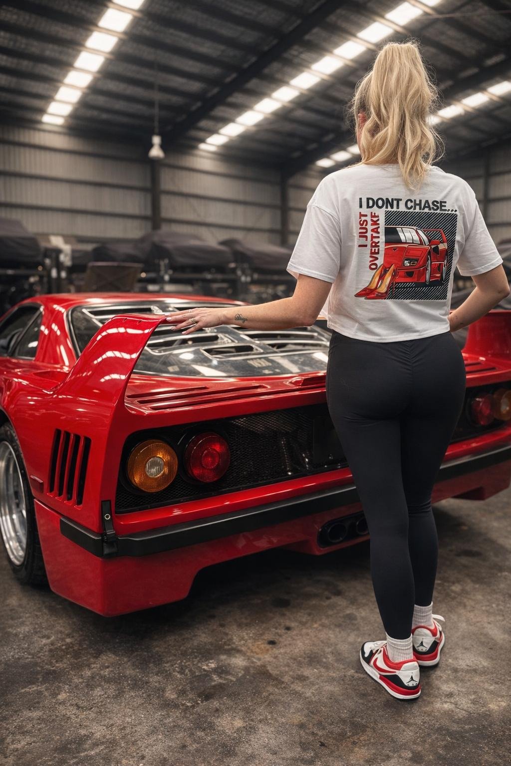A blonde women standing next to a Ferrari F40 car, whilst wearing a white crop top with the same Ferrari F40 graphic on the back back of the shirt with the text that states 'I dont chase, I just overtake' 
