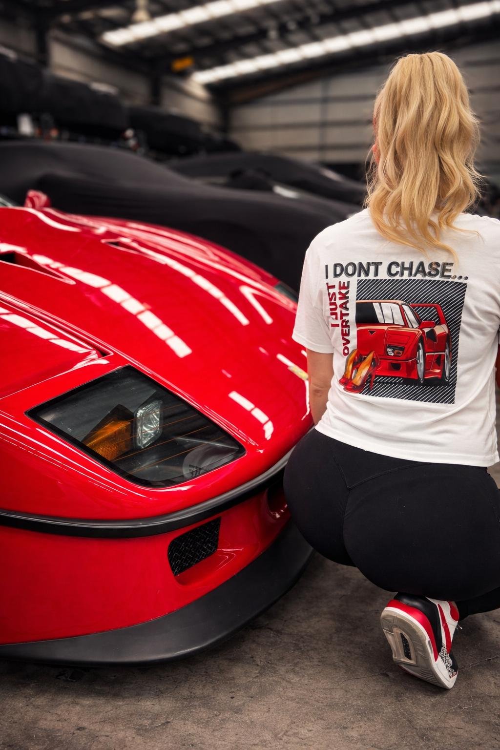 A woman with long blonde hair kneeling beside a red Ferrari F40 in a garage. She is wearing a white t-shirt with a red Ferrari F40 graphic and the words "I DON'T CHASE... I JUST OVERTAKE" printed on the back, black pants, and red and white sneakers.