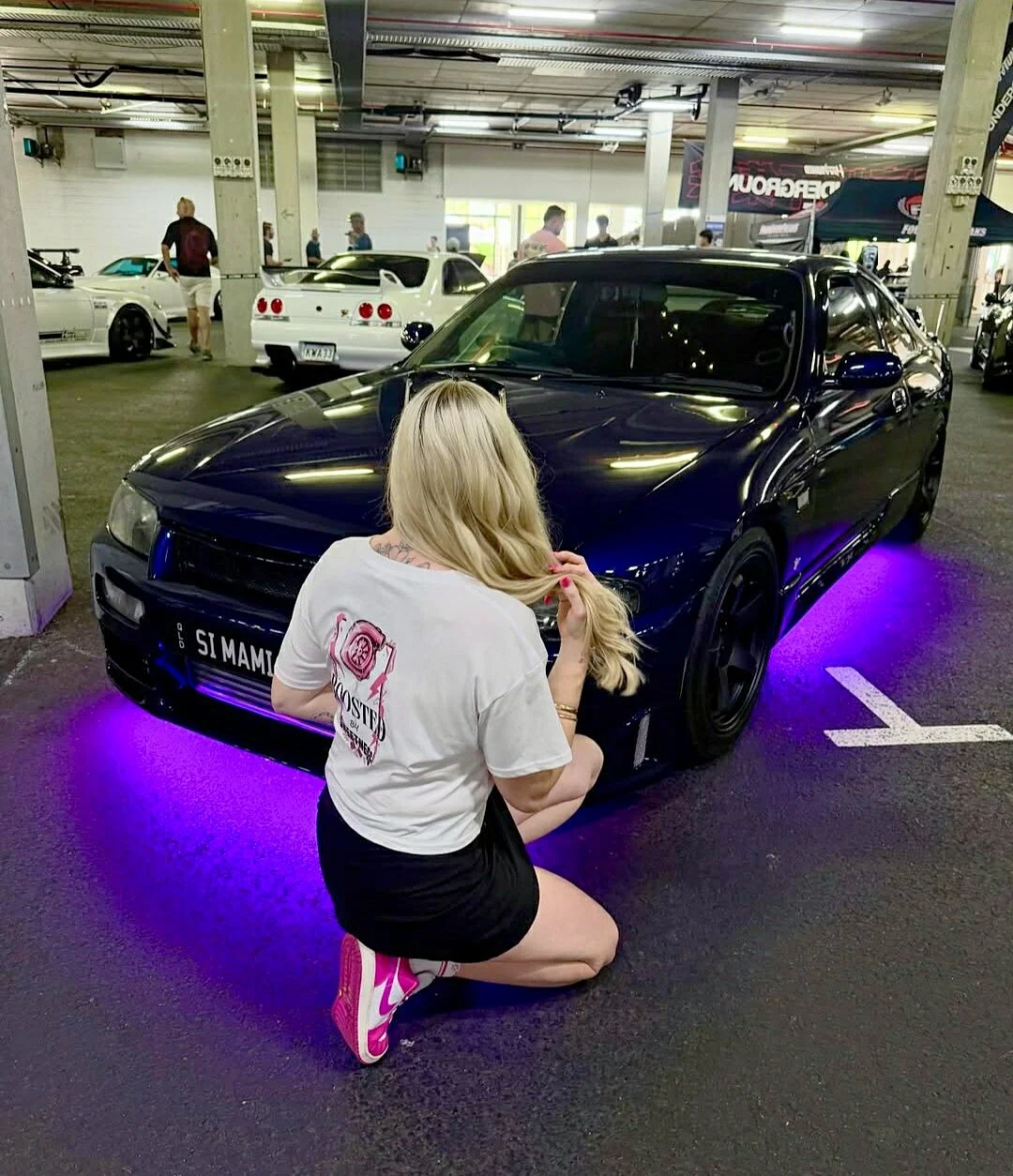 A young woman with blonde hair wearing white sunglasses, a white cropped t-shirt with a pink turbo graphic and text that says, 'Boosted by Sweetness', and black shorts, kneeling down in front of a Nissan R33 of an underground carpark. 