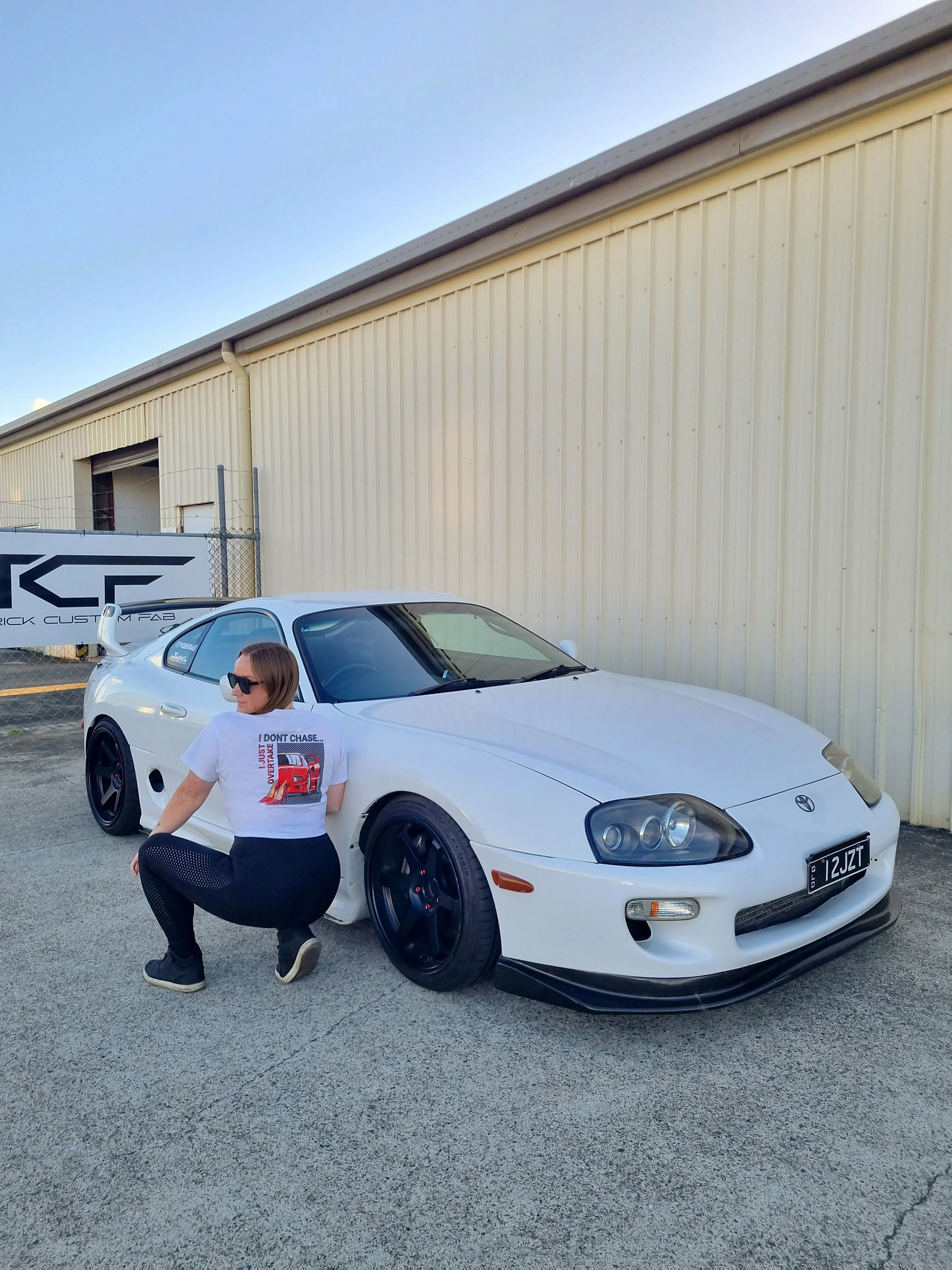 Back of a person wearing a white crop T-shirt with a graphic on the back, featuring an anime graphic of a young girl, with checkered background, and text 'I'm the Red Flag' under a cloudy sky crouching next to a white twin turbo with a 2JZ MK4 Supra.