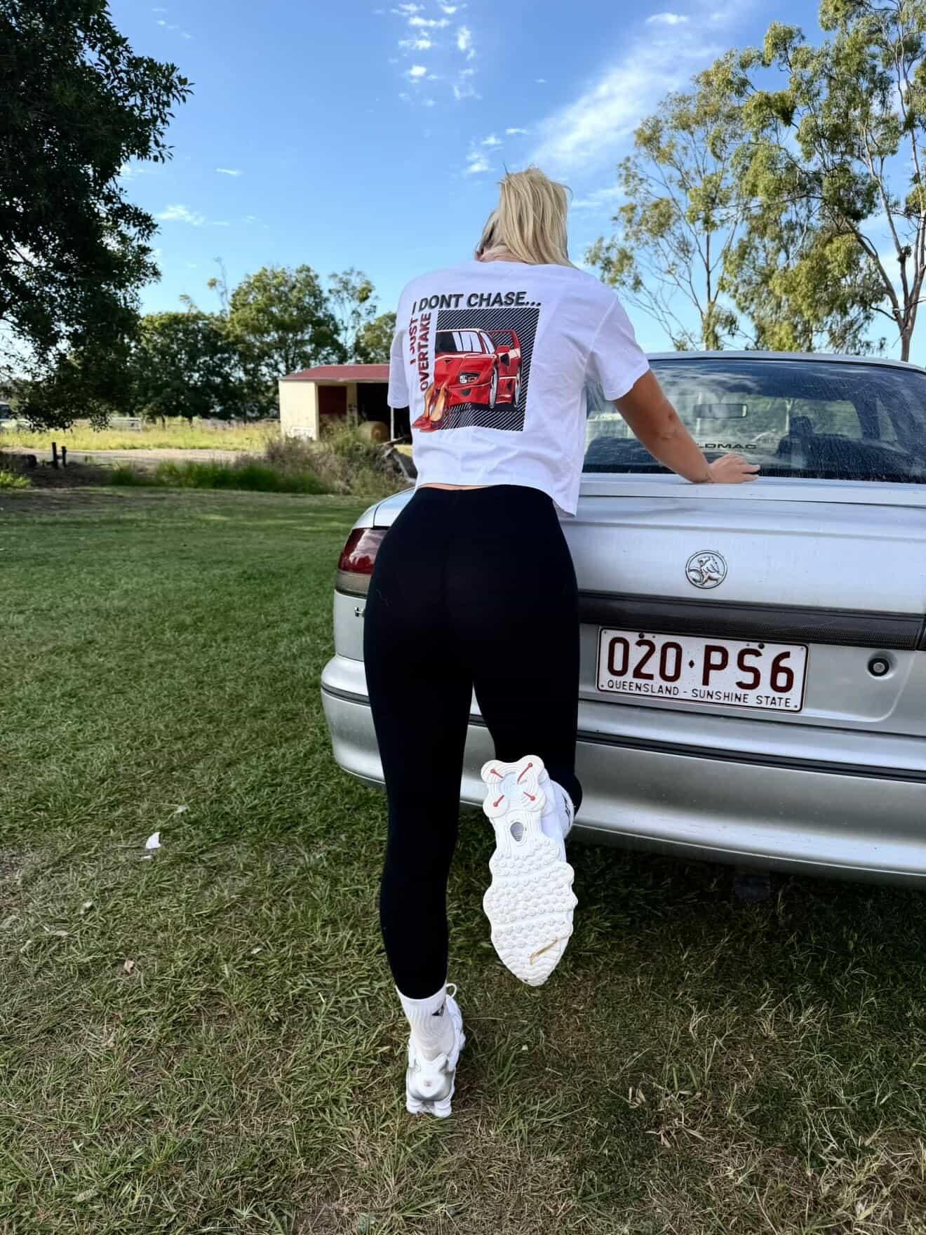 Back of a person with blonde hair up in a ponytail wearing a white crop T-shirt with a graphic on the back, which features an anime graphic of a young girl, with checkered background, and text 'I Dont Chase, I just Overtake' under a clear blue sky . 