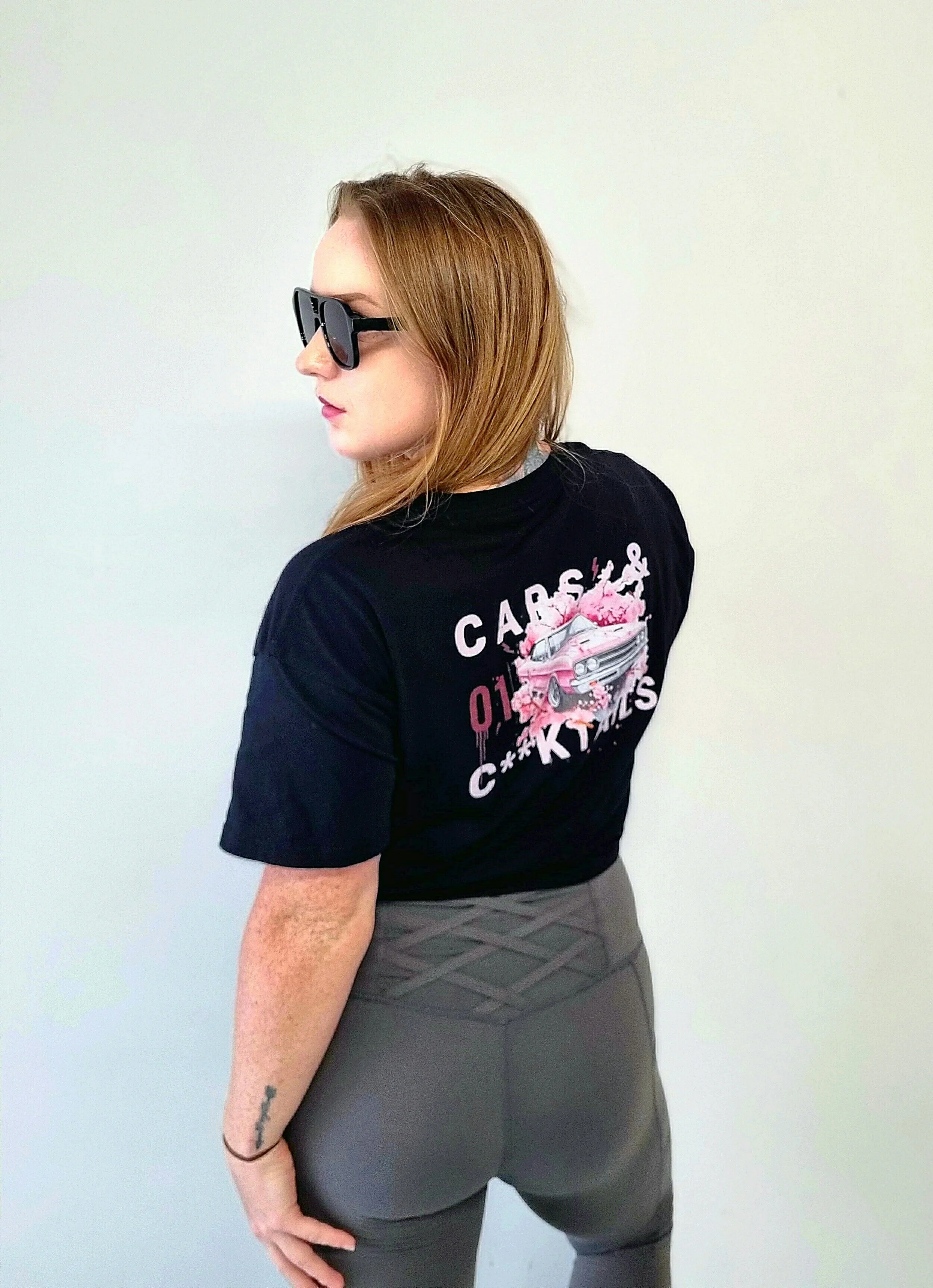 Young woman with light brown hair, wearing a black crop t-shirt and black designer glasses with a vintage car graphic and the text 'Cars and Cocktails' on her back, standing indoors against a crisp white wall with grey criss cross style leggings