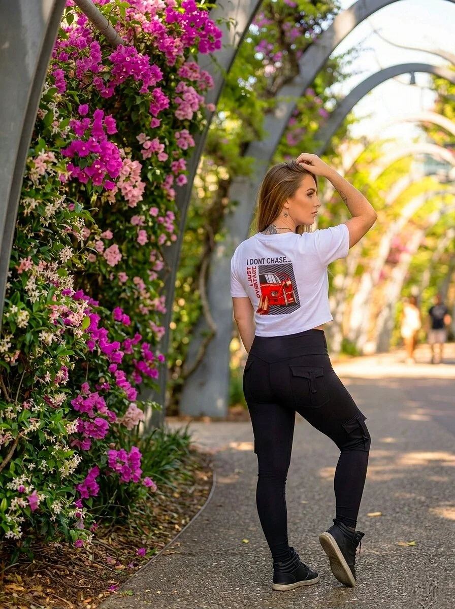 Young woman with brown hair, wearing a white crop t-shirt with a red sports car graphic and the text 'I DON'T CHASE...I JUST OVERTAKE' on her back, standing outdoors next to a colorful flower wall, with black cargo style pocket leggings and high ankl