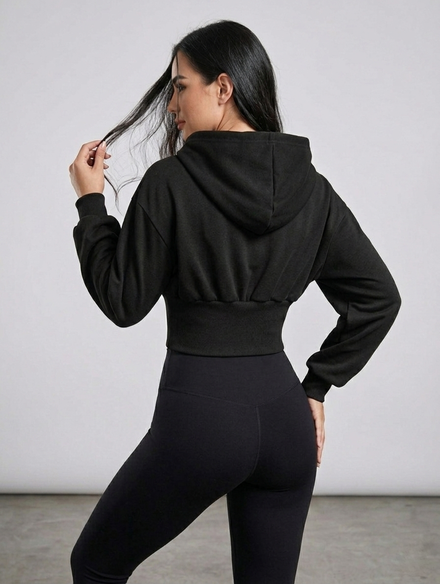 A woman standing in front of a crisp white wall playing with her black long hair, wearing a black cropped hoodie with a lace-up front and black high-waisted black leggings.
