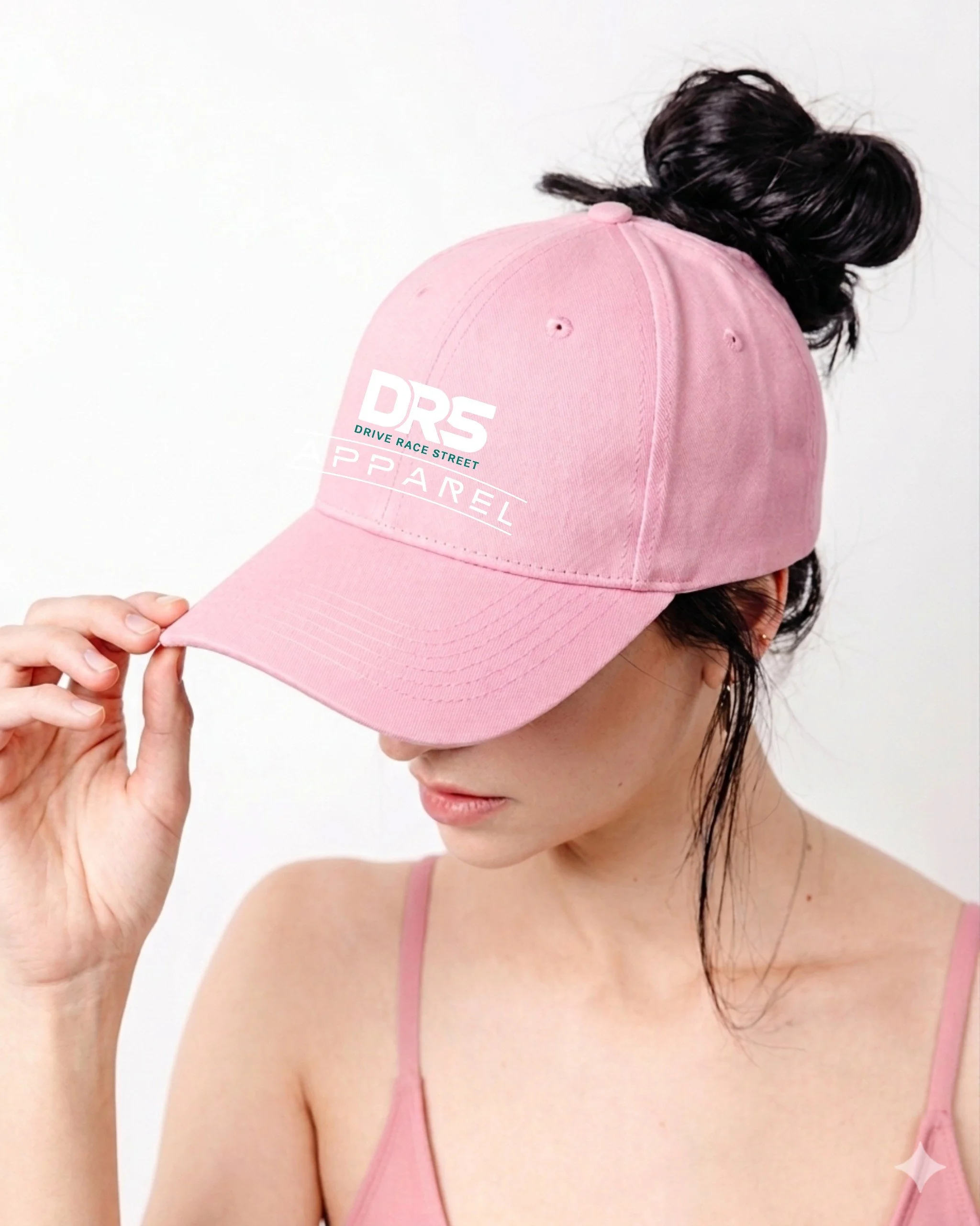 A women with brunette hair tied up in a bun, wearing a light pink high ponytail cap, with a magnetic back closure with a logo on the front from DRS Apparel.  