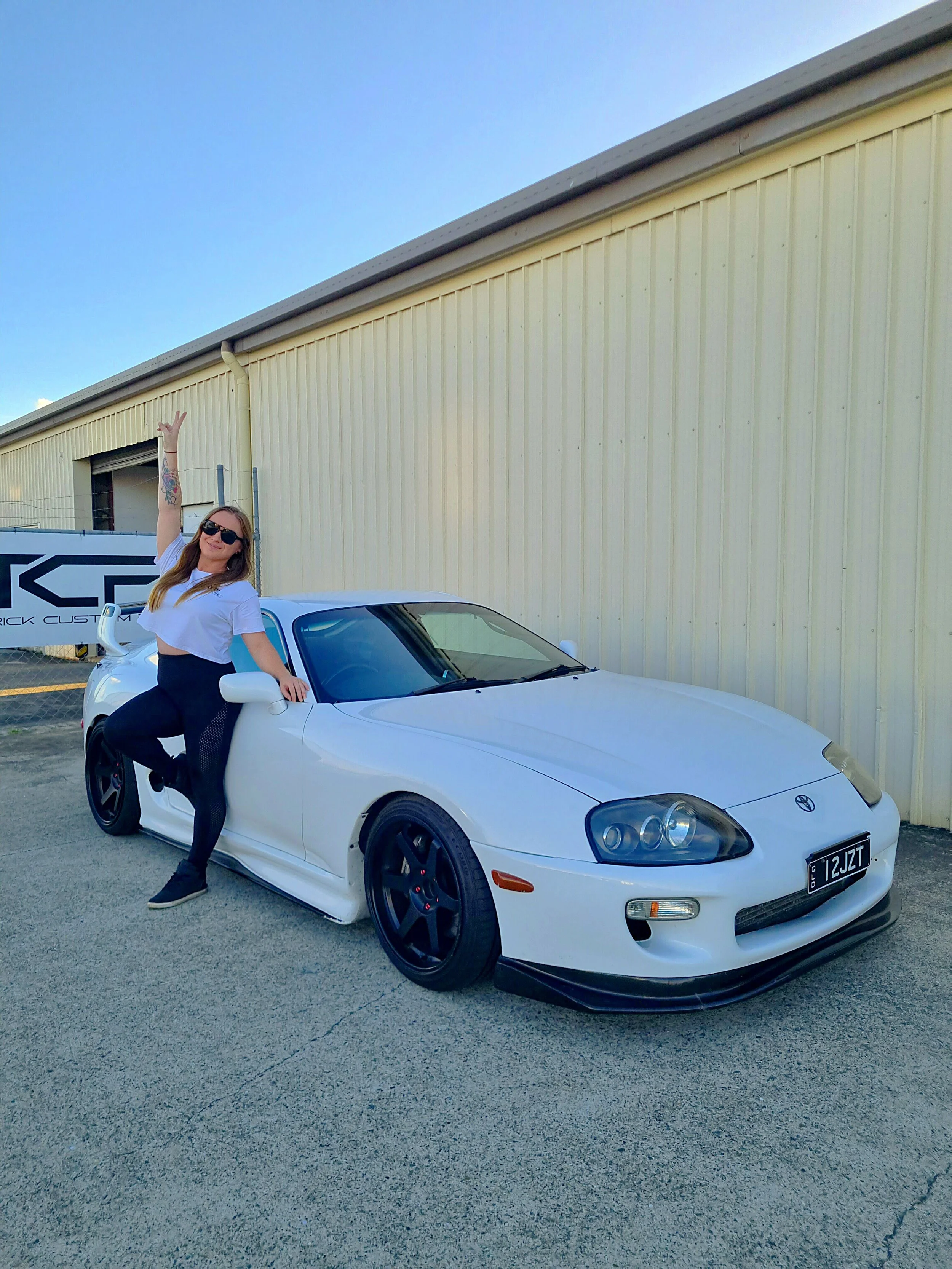 A woman with long hair, sunglasses, and tattoos on her right arm, dressed in a white crop top and black pants, is posing next to a white MK4 Supra with black TE37 wheels, parked outside a beige industrial building of Tyre and Wheel Solutions.