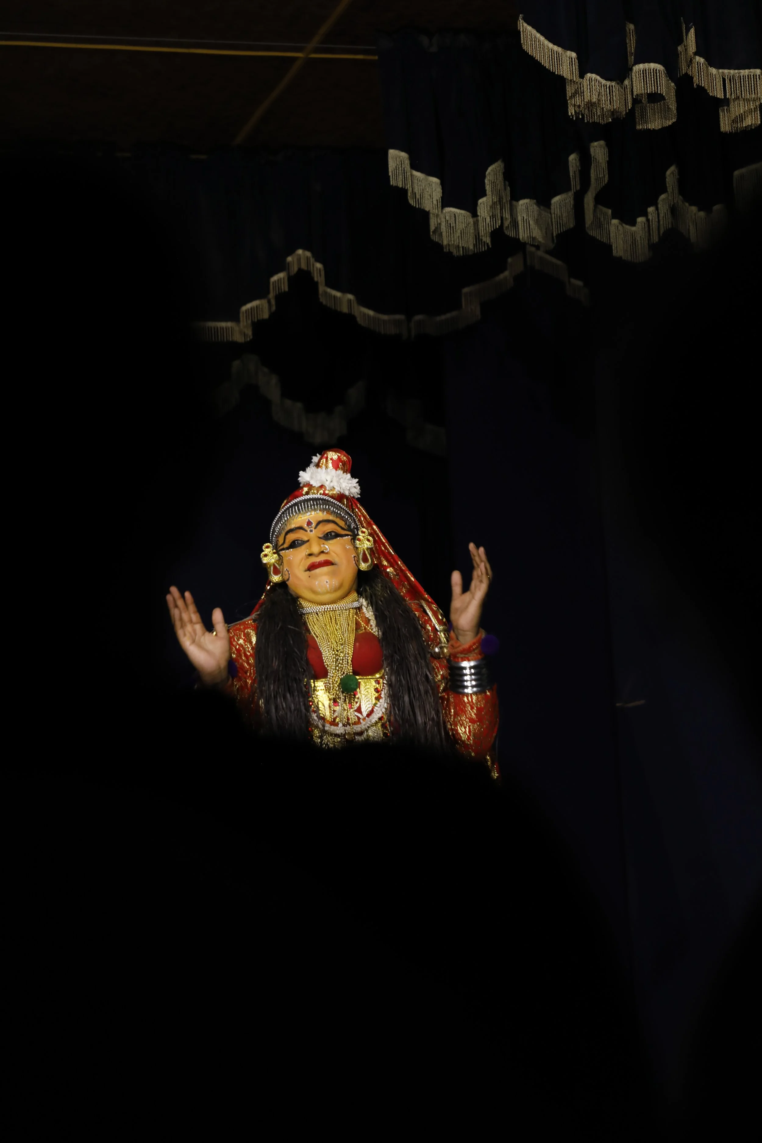 A traditional Indian puppet dressed in a red and gold costume with jewelry, performing on stage with dark curtains in the background.