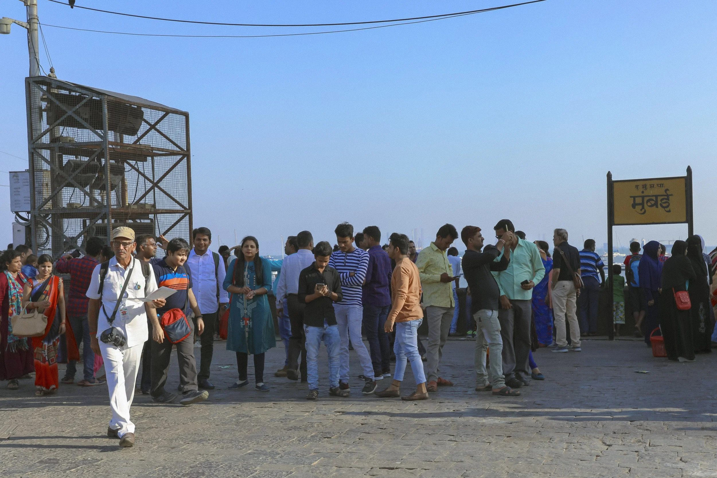 A crowd of people gathers near a dock in Mumbra, with some individuals engaged on their phones and others walking or standing around. A sign reads "Mumbra" in Hindi and English. The background shows a clear blue sky and some boats docked in the water