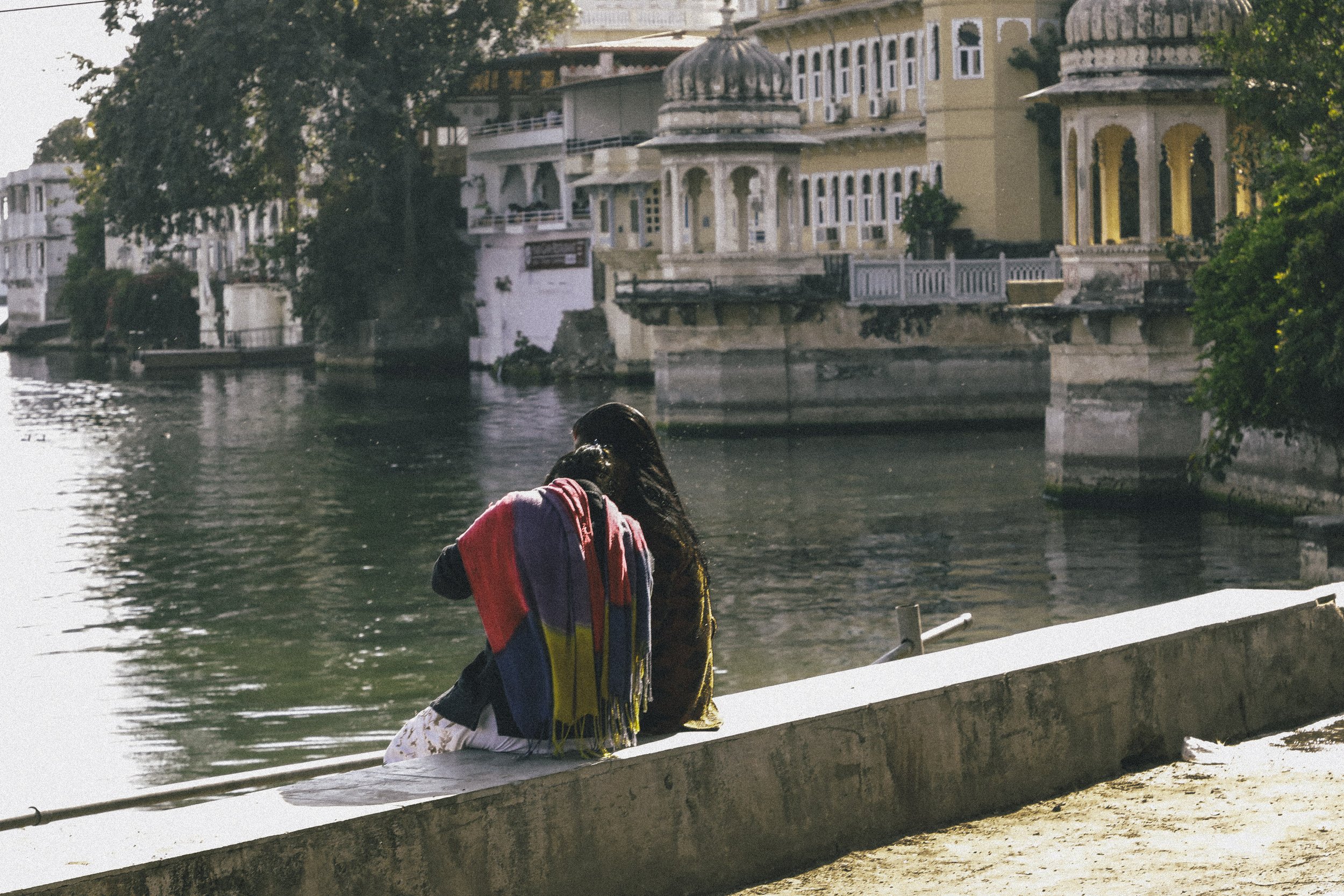 Two women sitting by a river, one wearing a striped shawl and the other with long black hair, with a backdrop of ornate buildings and greenery.