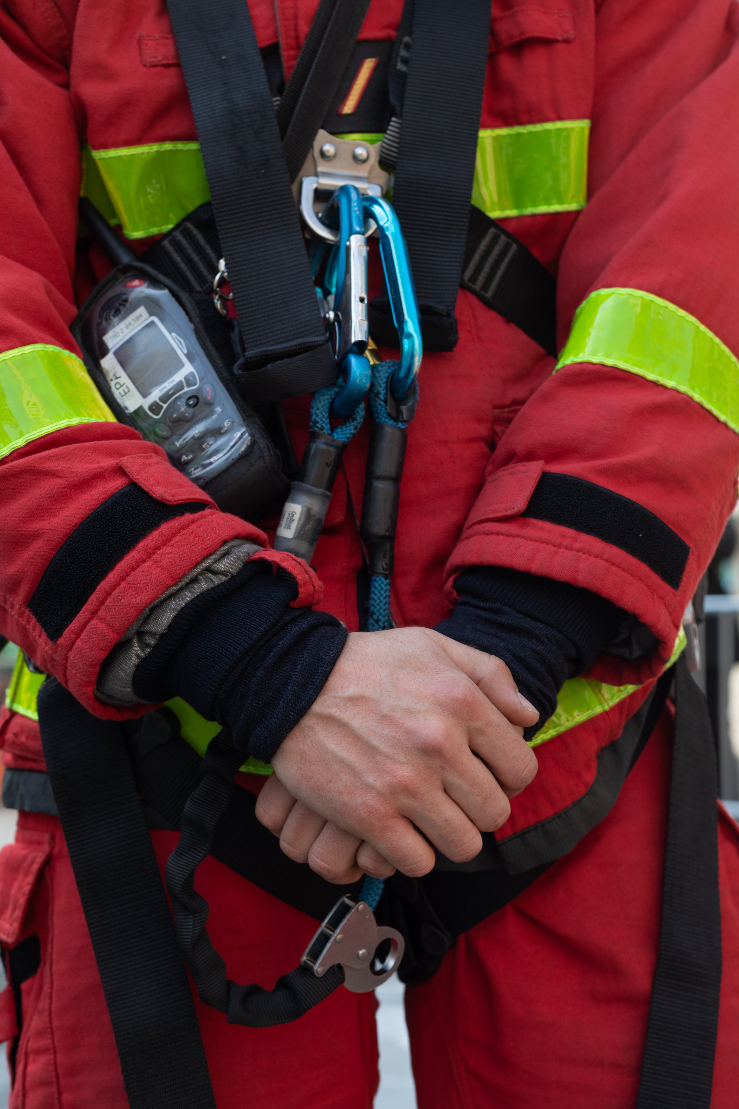 Close-up of a person in a red safety uniform with reflective yellow stripes, wearing a harness with carabiners and safety equipment, and holding their hands together.