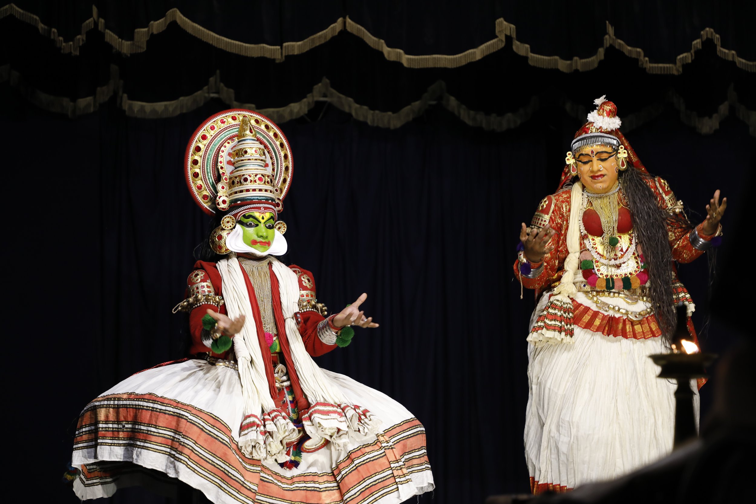 Two performers dressed in traditional Indian Kathakali costumes on stage. The performer on the left has green face paint and elaborate headgear, sitting cross-legged. The performer on the right has detailed makeup, long hair, and is standing with han