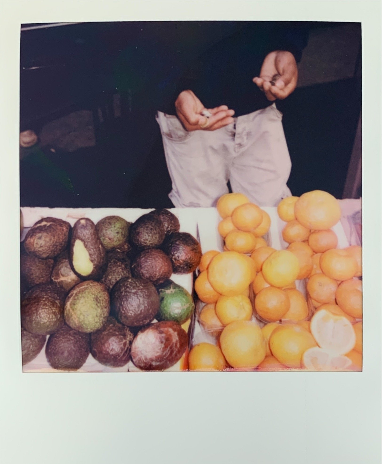 A person standing behind a table of fresh avocados and oranges at a market stall, with their hands making a gesture.