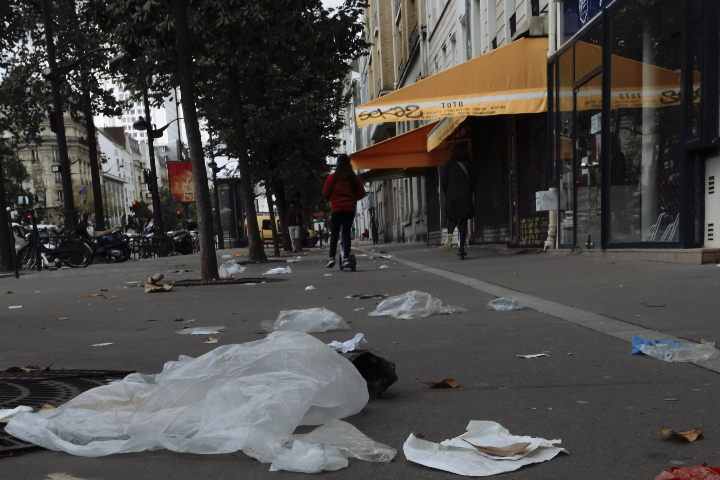 Empty city sidewalk littered with trash and plastic bags, with a person riding a scooter and people walking, storefronts with umbrellas, and trees lining the street.