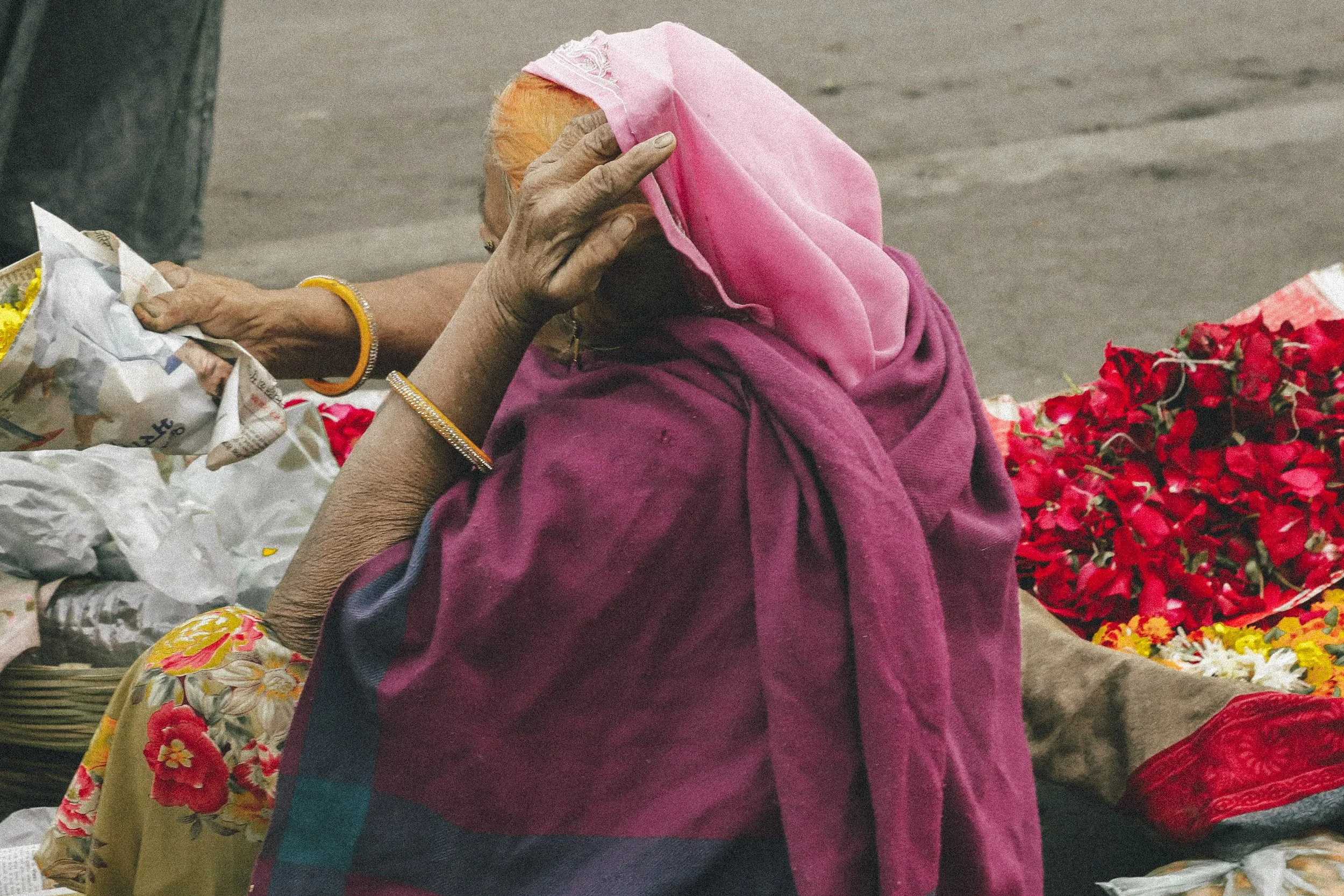 An elderly woman wearing a pink headscarf and a maroon shawl, sitting on the ground surrounded by flowers and vendors.