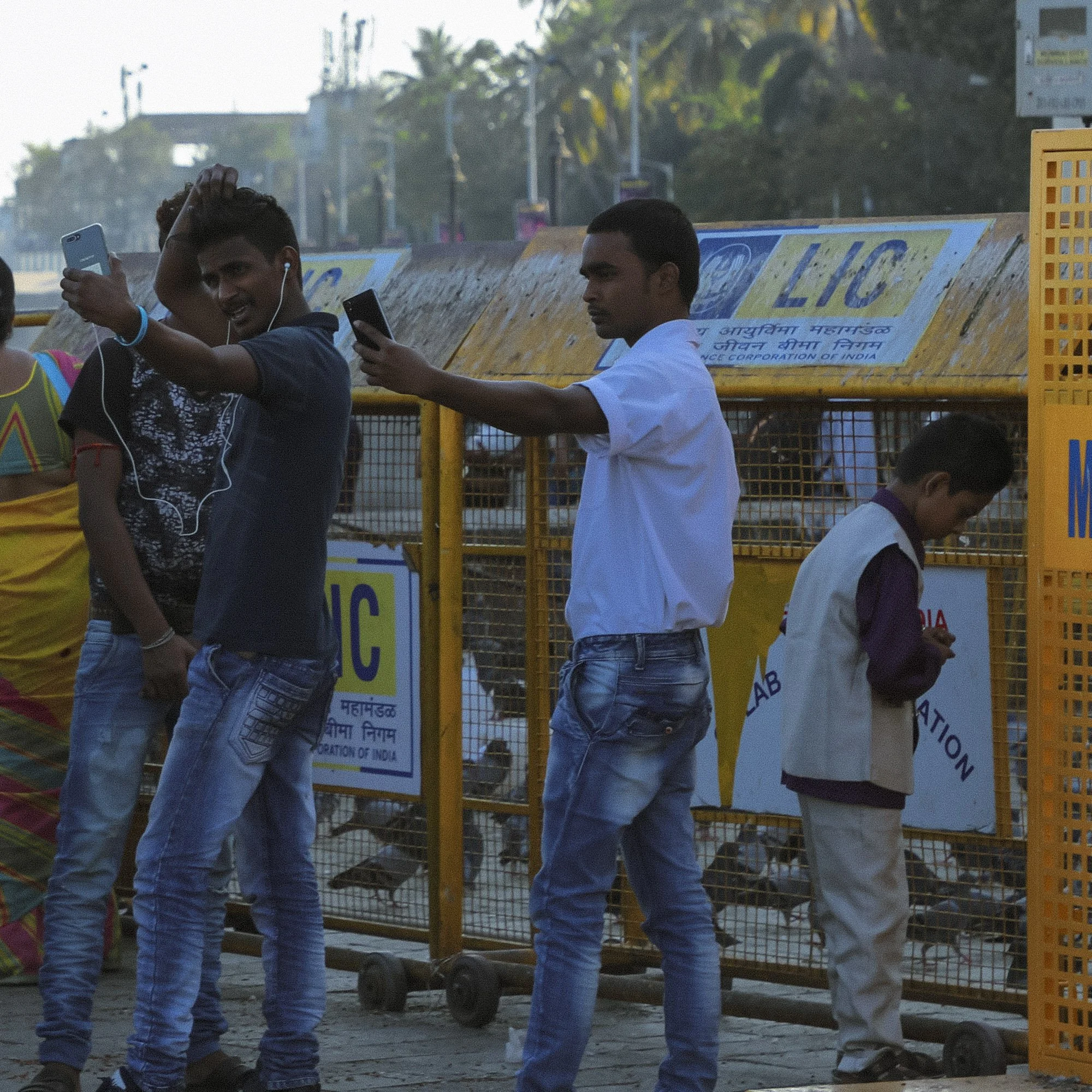 Three young men standing near a yellow fence with licenses sign, taking selfies with their phones, while a child stands nearby.