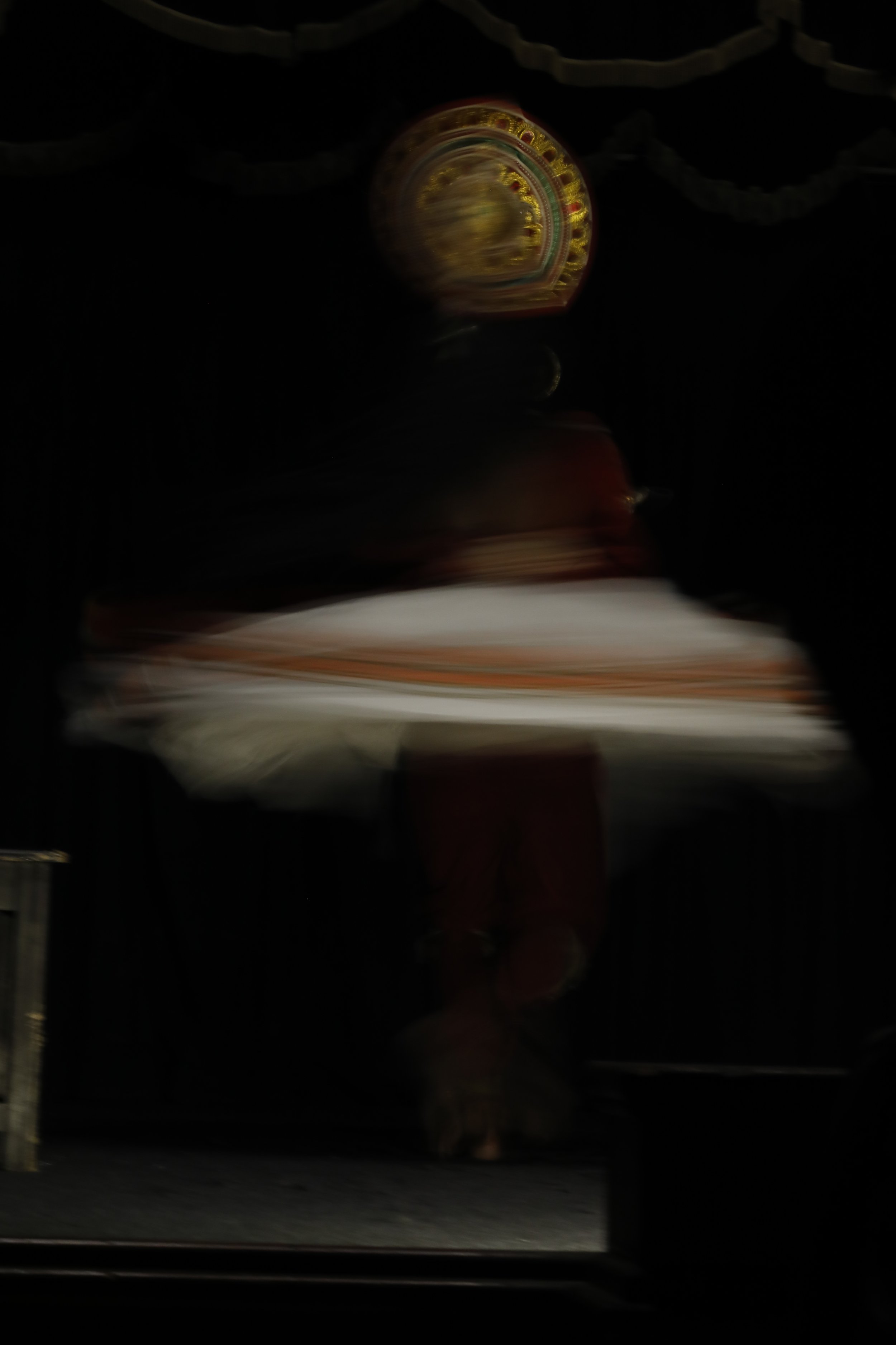 A blurred image of a person wearing traditional attire on a stage with decorative black and gold drapes in the background.