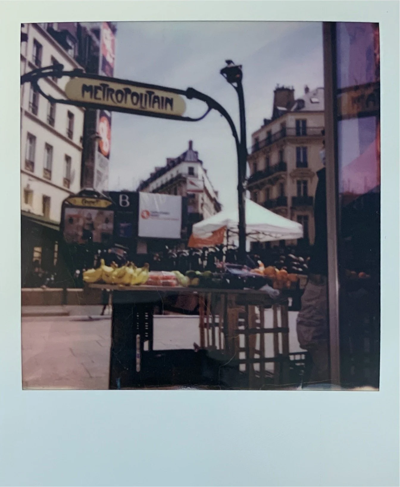 Street scene in Paris with a 'Metropolitain' subway sign, outdoor market stall with bananas and other fruits, historic buildings, and a white tent in the background.