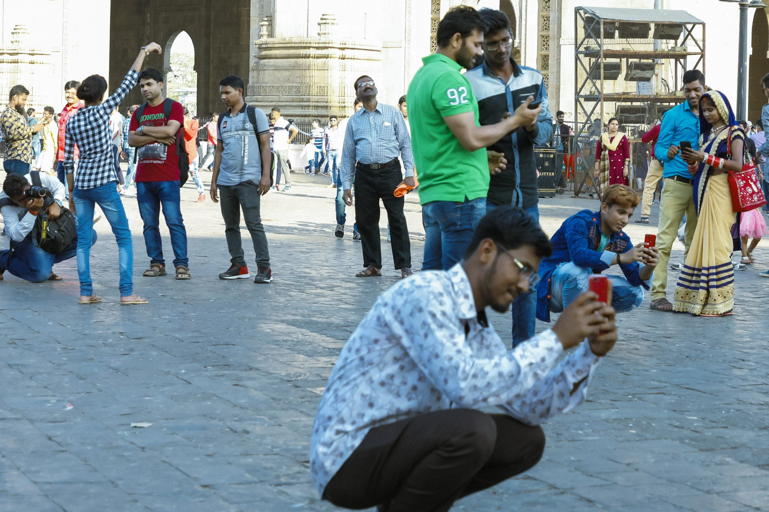 Group of people, mostly young men and women, taking photos and selfies outside a historical monument in India, with some wearing traditional attire and others in casual clothing.