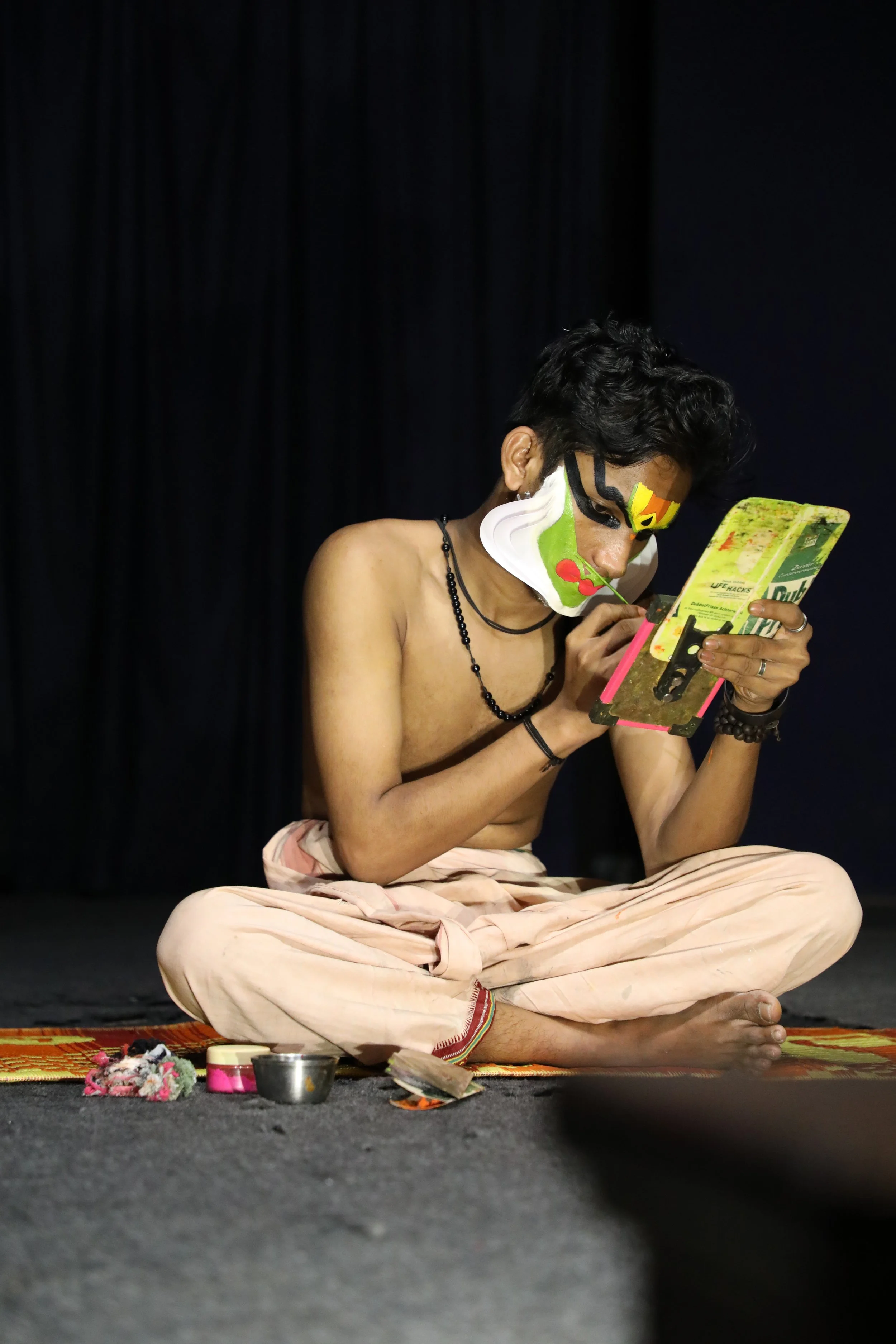 Person sitting cross-legged on stage, shirtless, wearing light-colored pants, with painted face, looking at a mirror, surrounded by small objects.