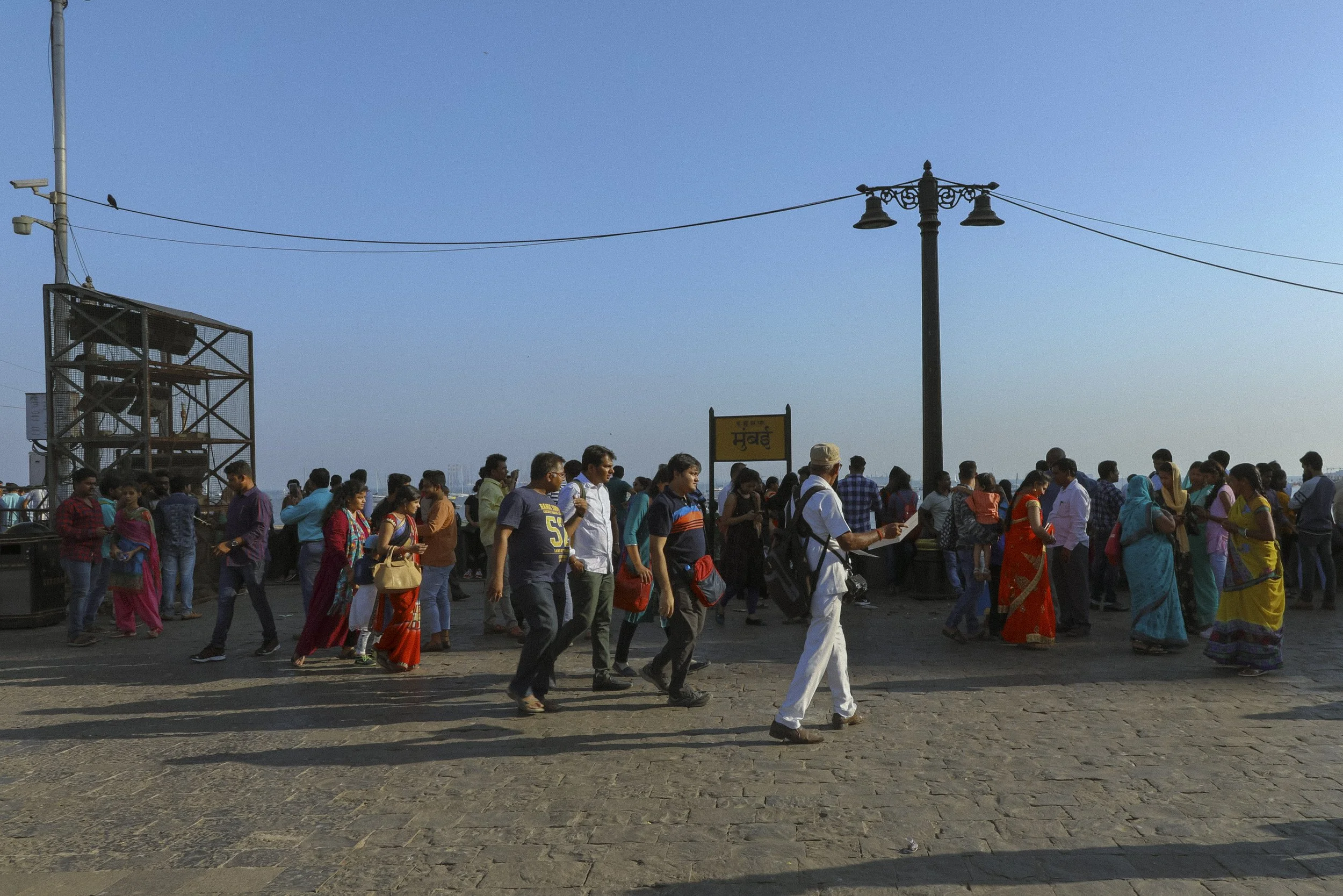 Crowd of people walking outdoors near a yellow sign with text in Hindi and English, under a clear blue sky, with streetlights and electrical wires overhead.
