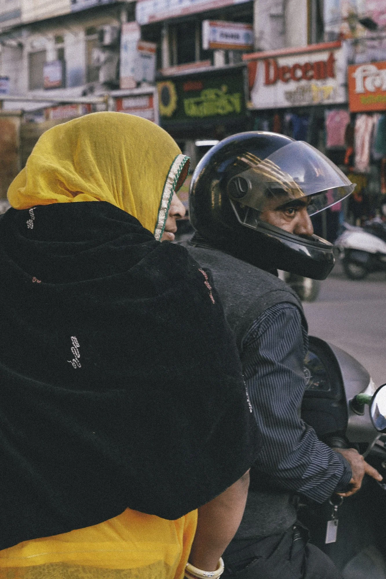 A woman with a yellow headscarf standing close behind a man in a motorcycle helmet riding a scooter on a city street, with various signs and buildings in the background.