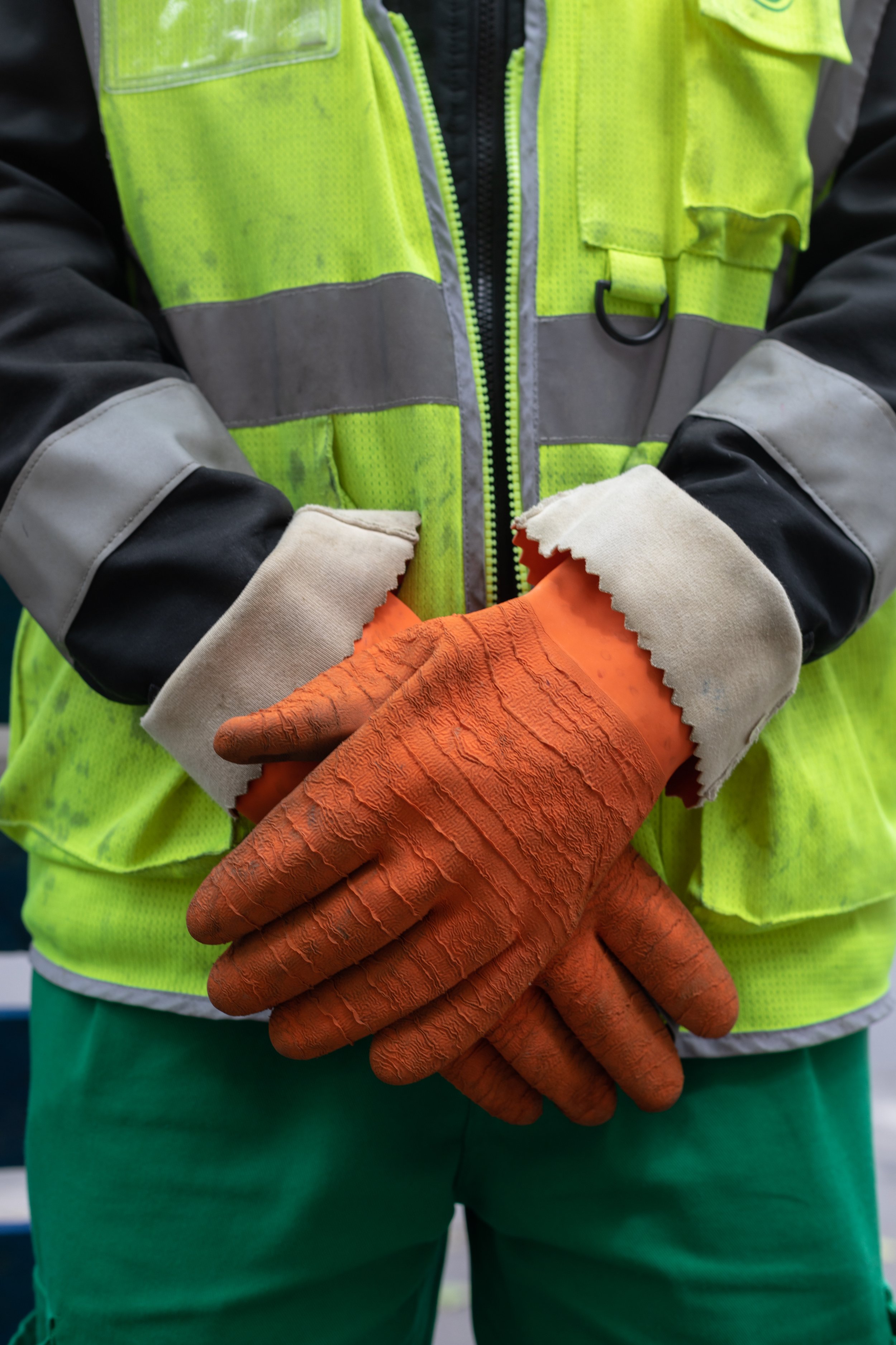 Close-up of a person wearing a neon yellow safety vest with gray and black sleeves, and orange textured work gloves, standing with their hands clasped.
