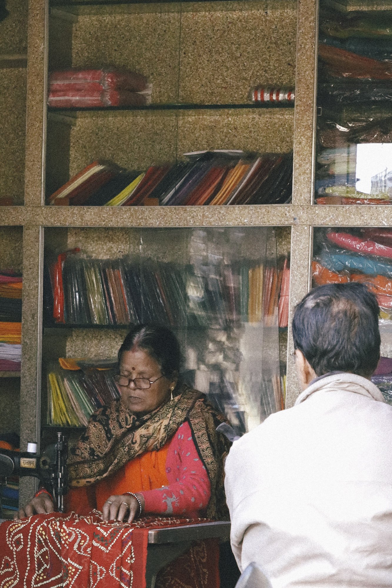 An elderly woman with glasses seated at a sewing machine, working on a red fabric with white dotted patterns, in front of a glass window with shelves filled with colorful fabric and files.